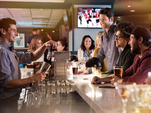 A bartender serving drinks to customers at a lively bar with sports playing on TVs.