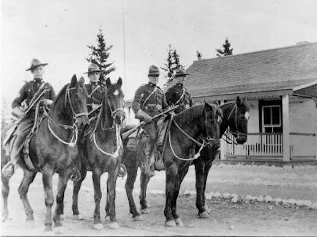 Four mounted police officers in uniform pose on horseback in front of a log building with a covered porch.