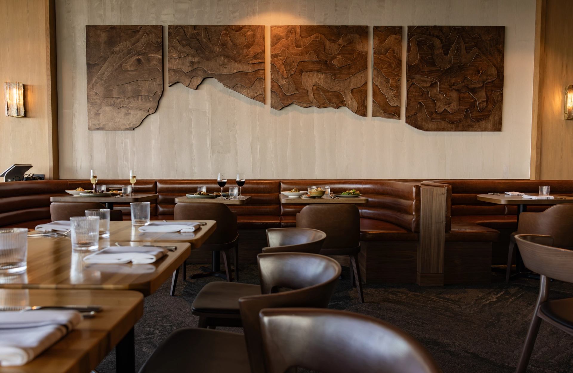 Restaurant dining room with brown leather banquettes, set tables and abstract wooden wall art.