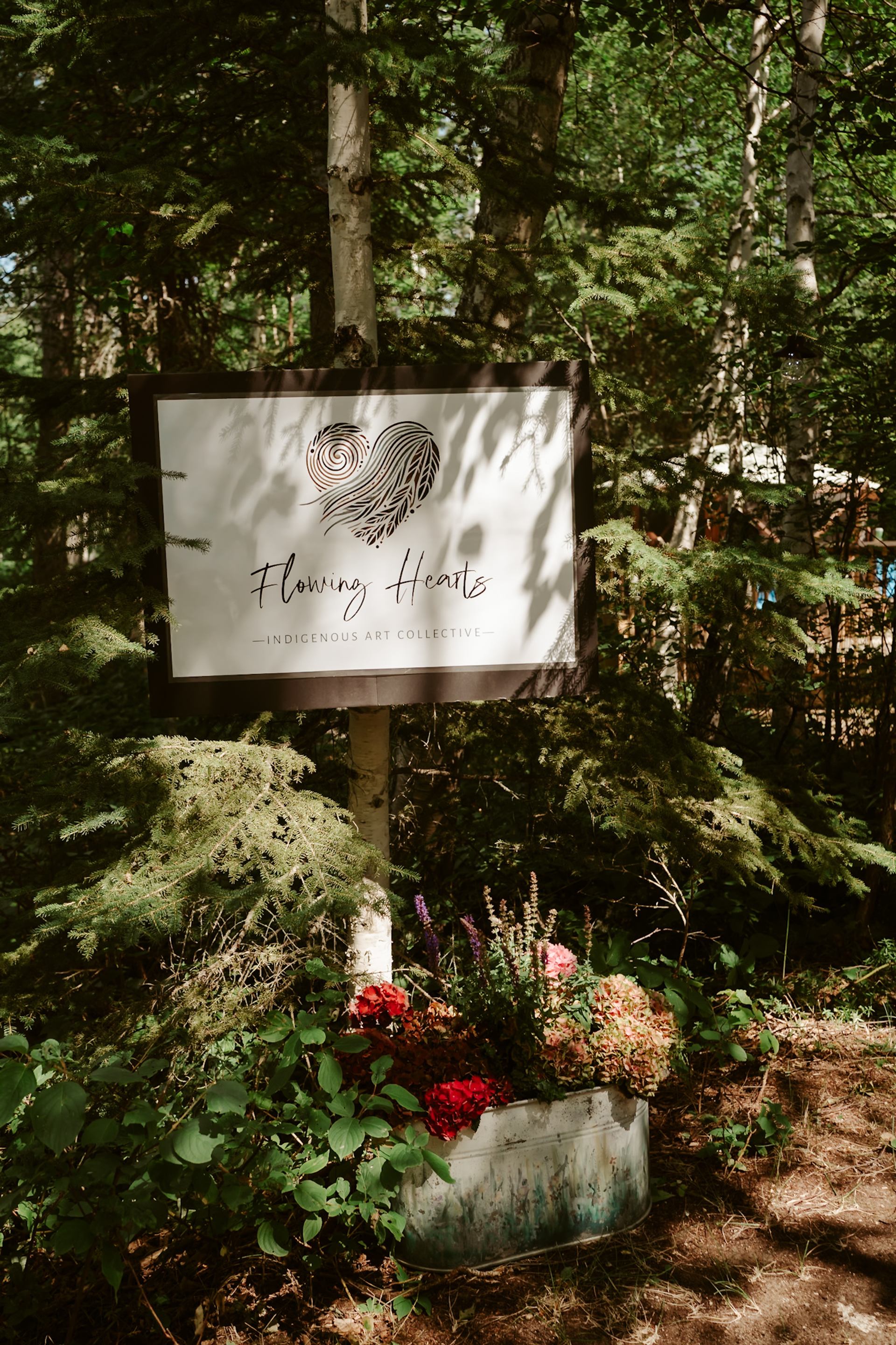 Flowing Hearts Indigenous Art Collective sign among trees and flowers.