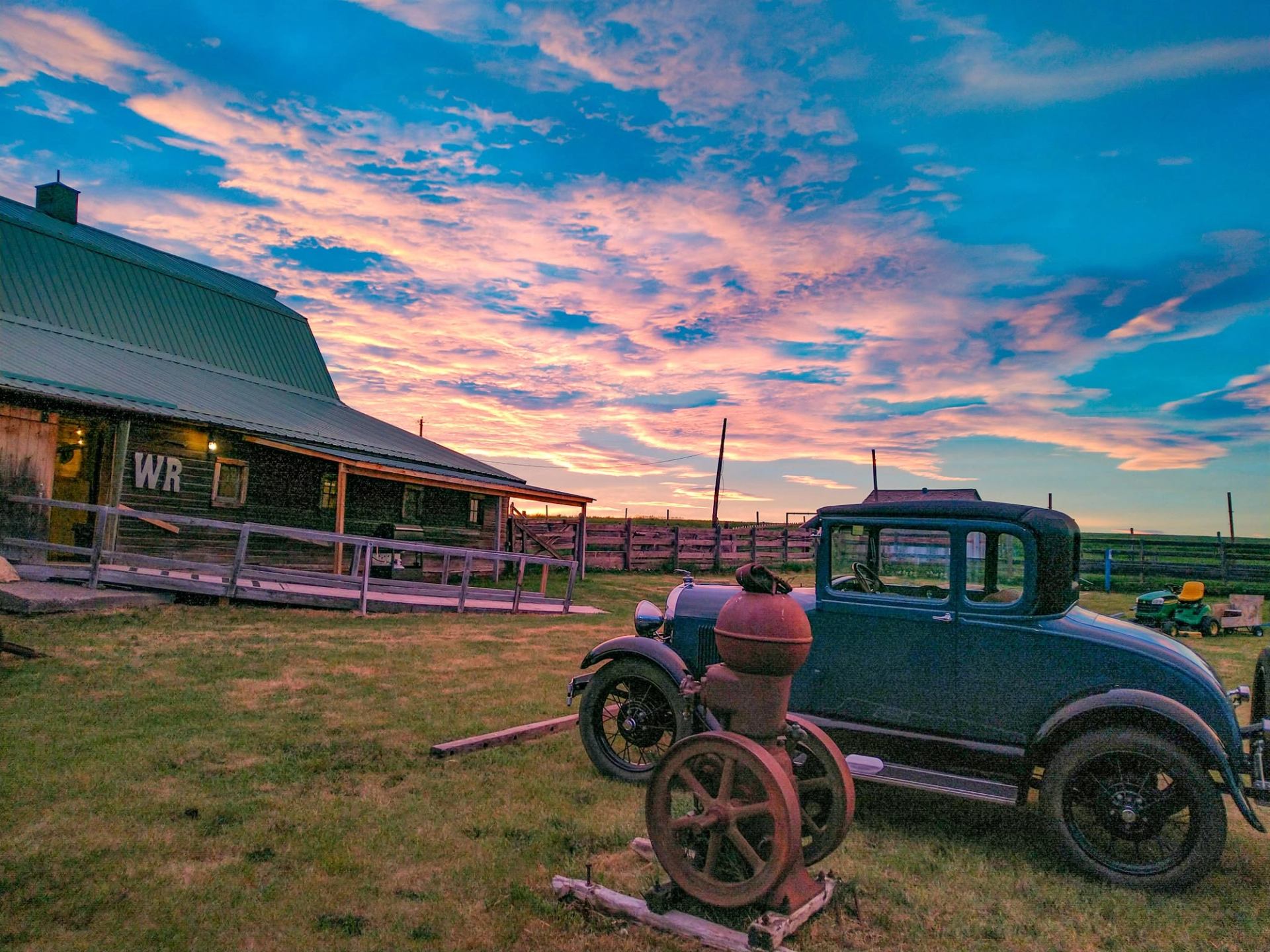 Vintage car and old farm equipment beside barn under colourful sunset sky.