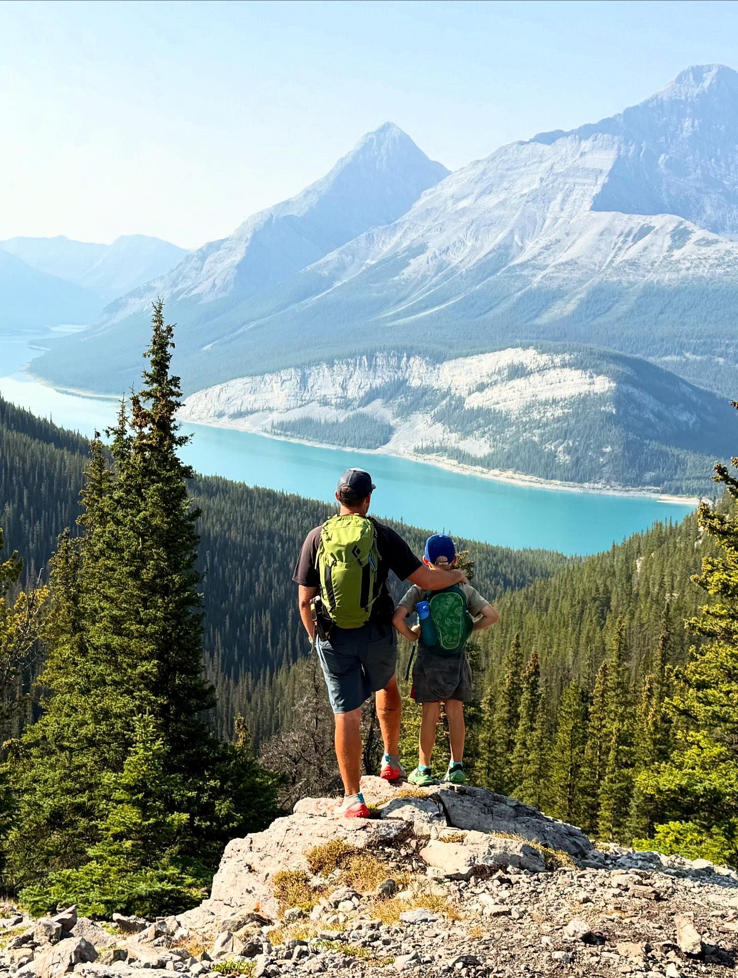 Two hikers with backpacks stand on rocky lookout above turquoise mountain lake.