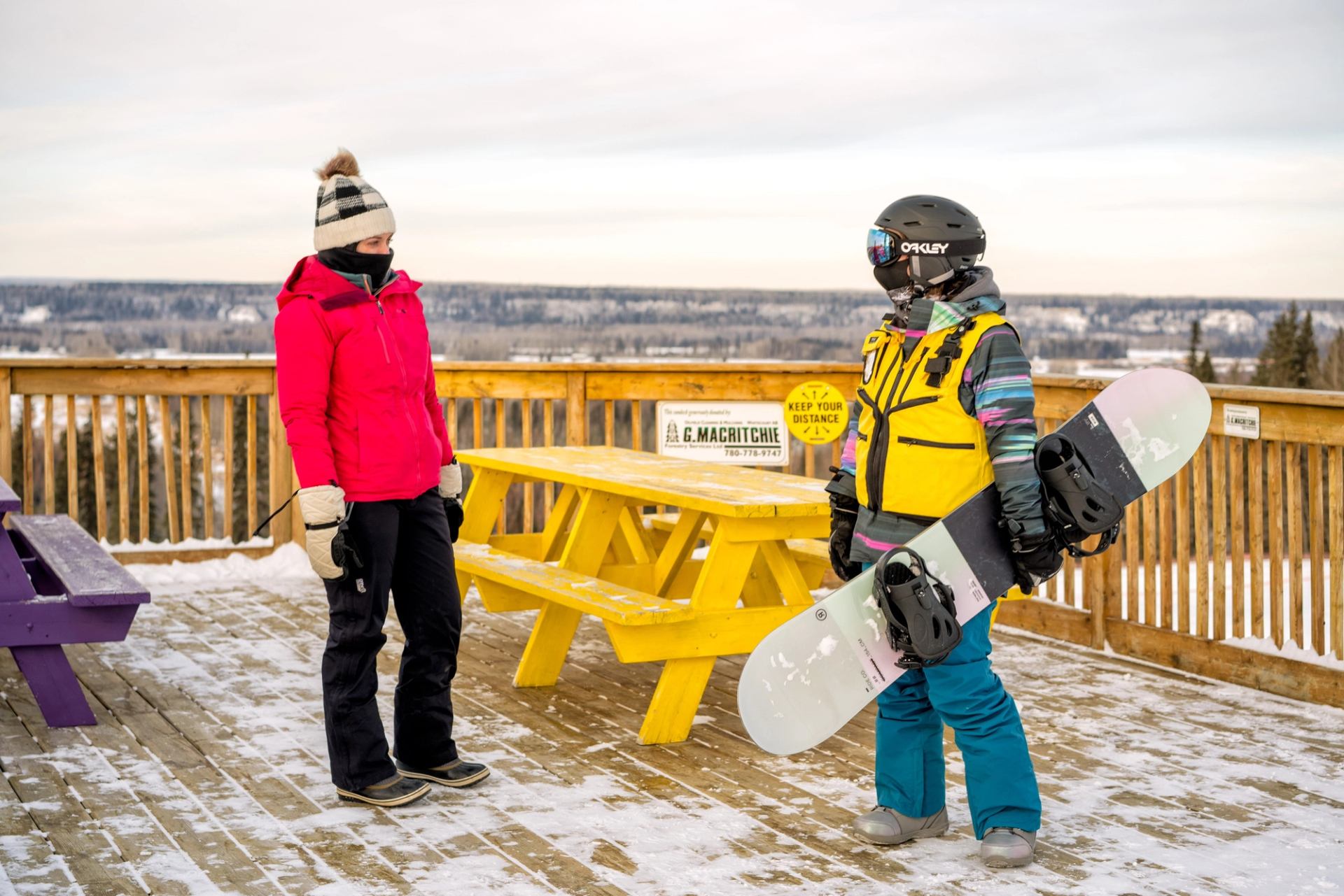 Two people in winter gear stand on snowy deck with a yellow table and snowboard.
