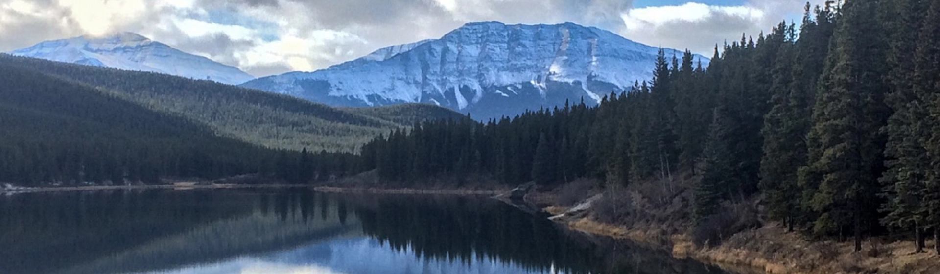 Snow-capped mountains reflected in a calm lake surrounded by dense evergreen forest.