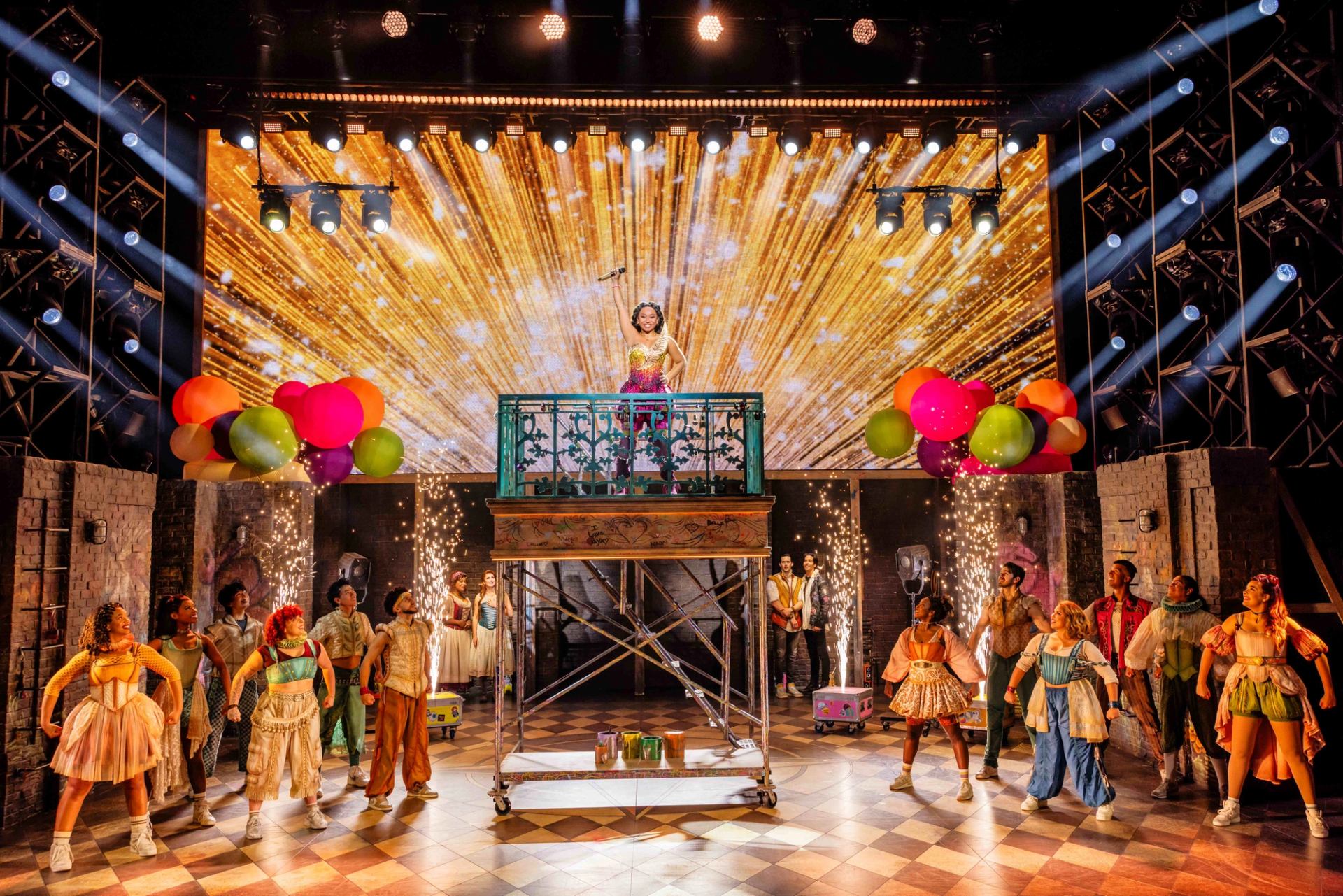 Stage musical cast surrounds raised platform with performer, balloons and golden fireworks backdrop.