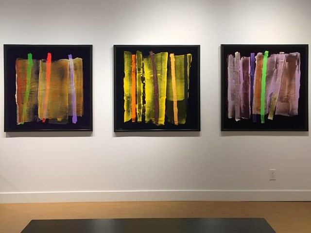 Three framed abstract paintings with vertical colour bands on a white gallery wall.