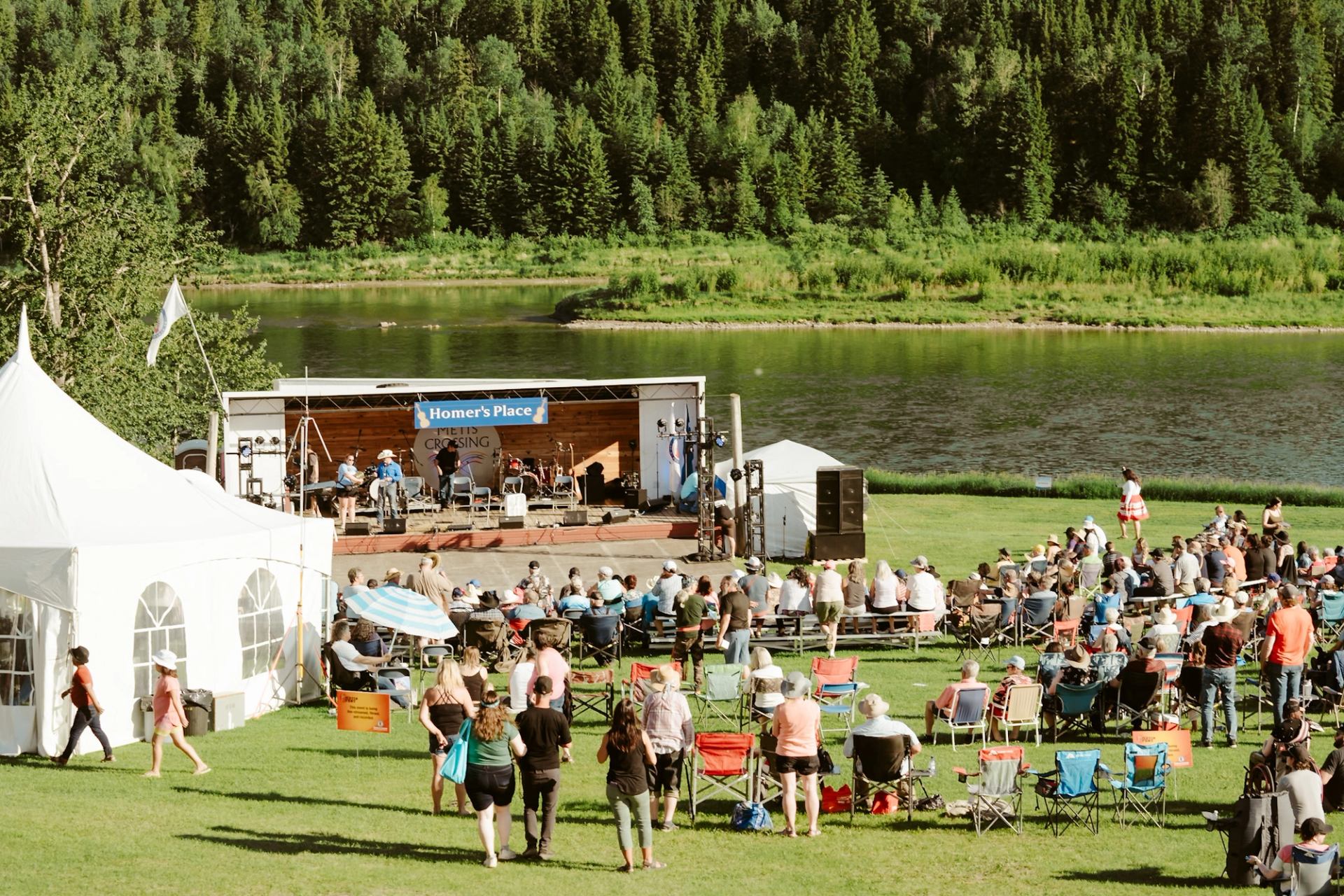 Outdoor riverside concert at Homer’s Place with seated crowd and forested backdrop.