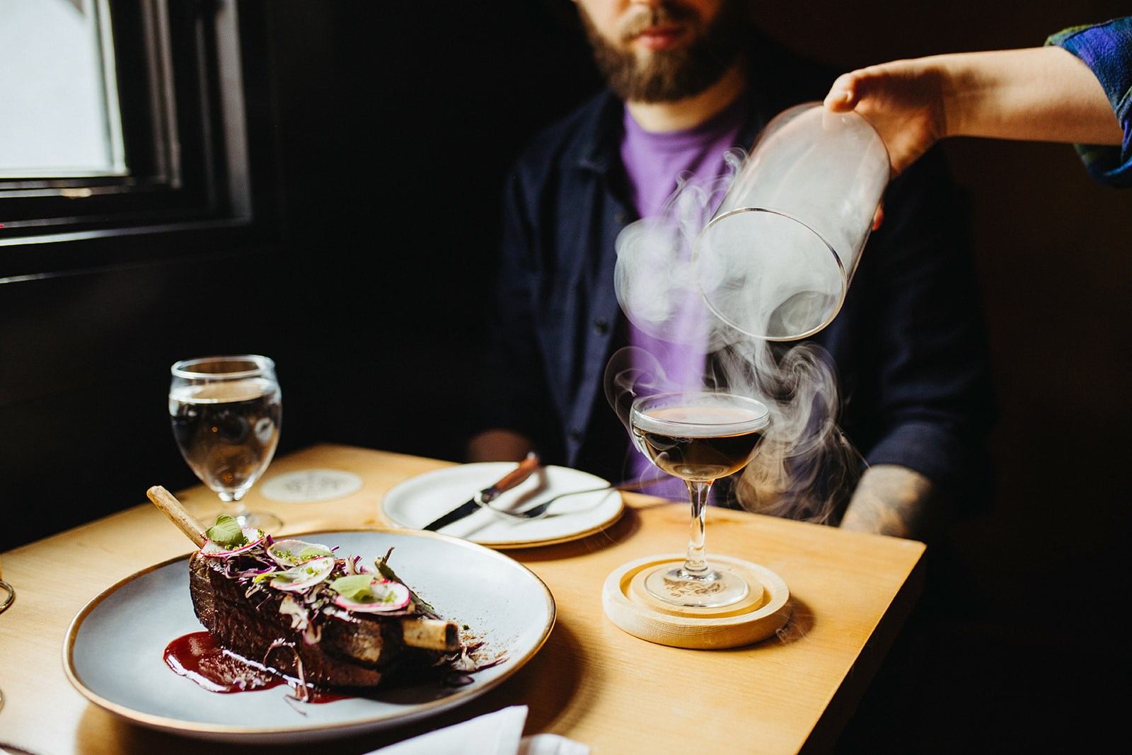 A smoky cocktail being poured over a dish of food at a dimly lit restaurant.