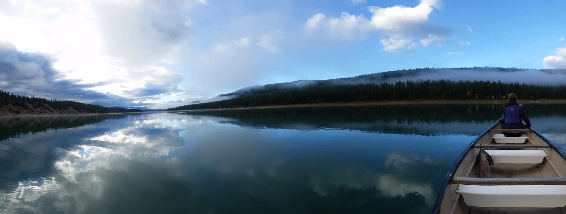 Canoe gliding across a calm lake with reflections of clouds and forested hills.