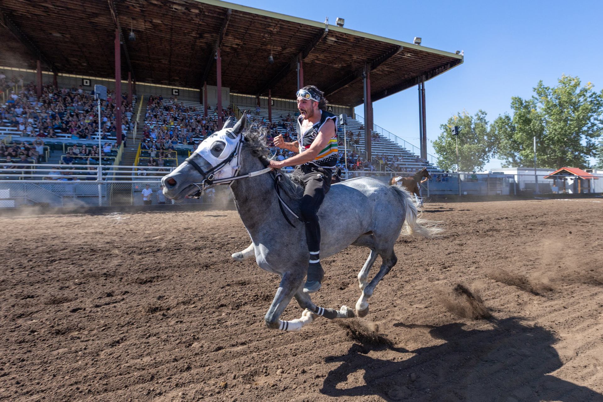 Bareback rider on galloping horse in dirt arena with grandstand crowd in background.