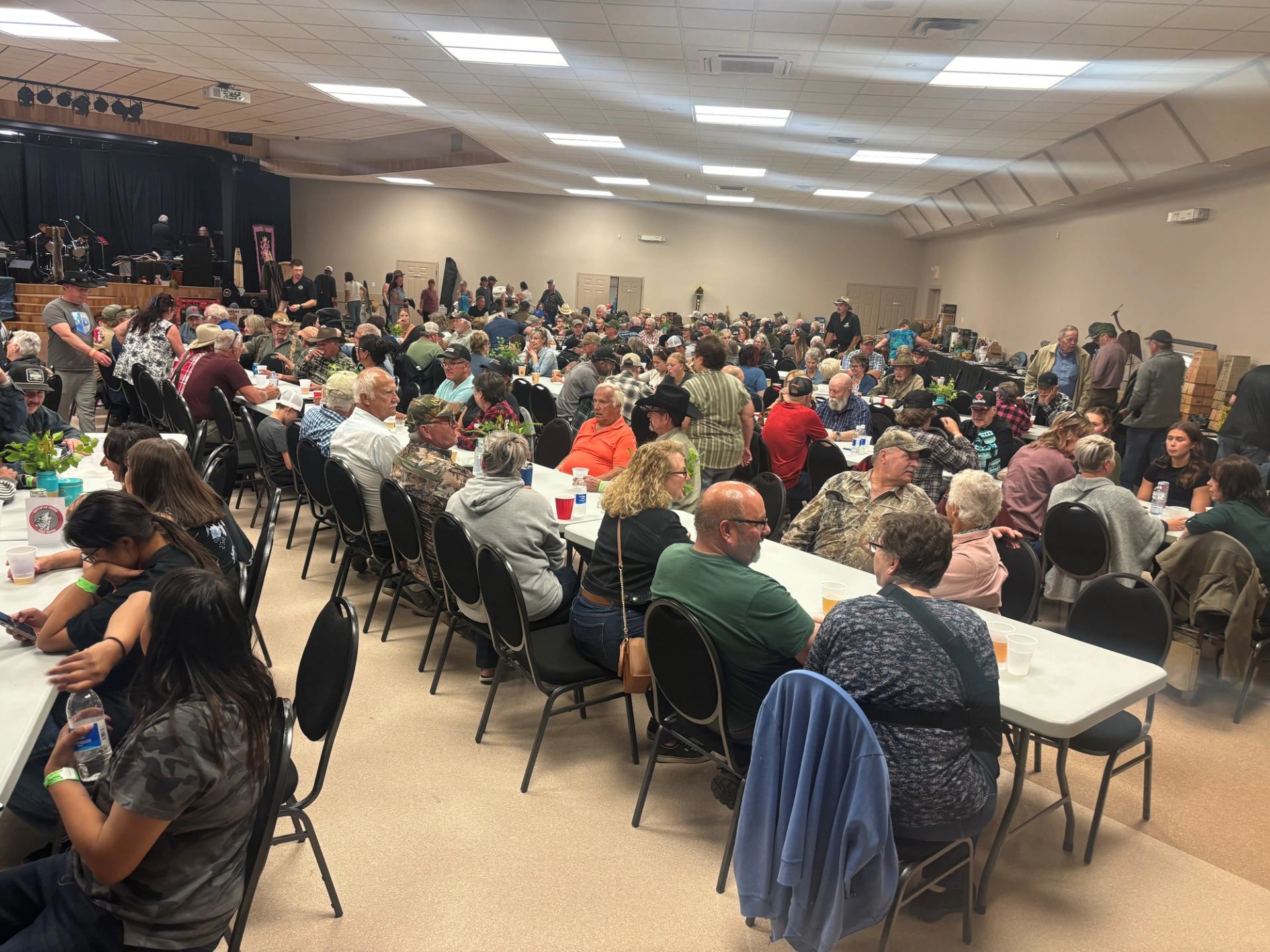 Crowded community hall with people seated at long tables, talking and eating together.