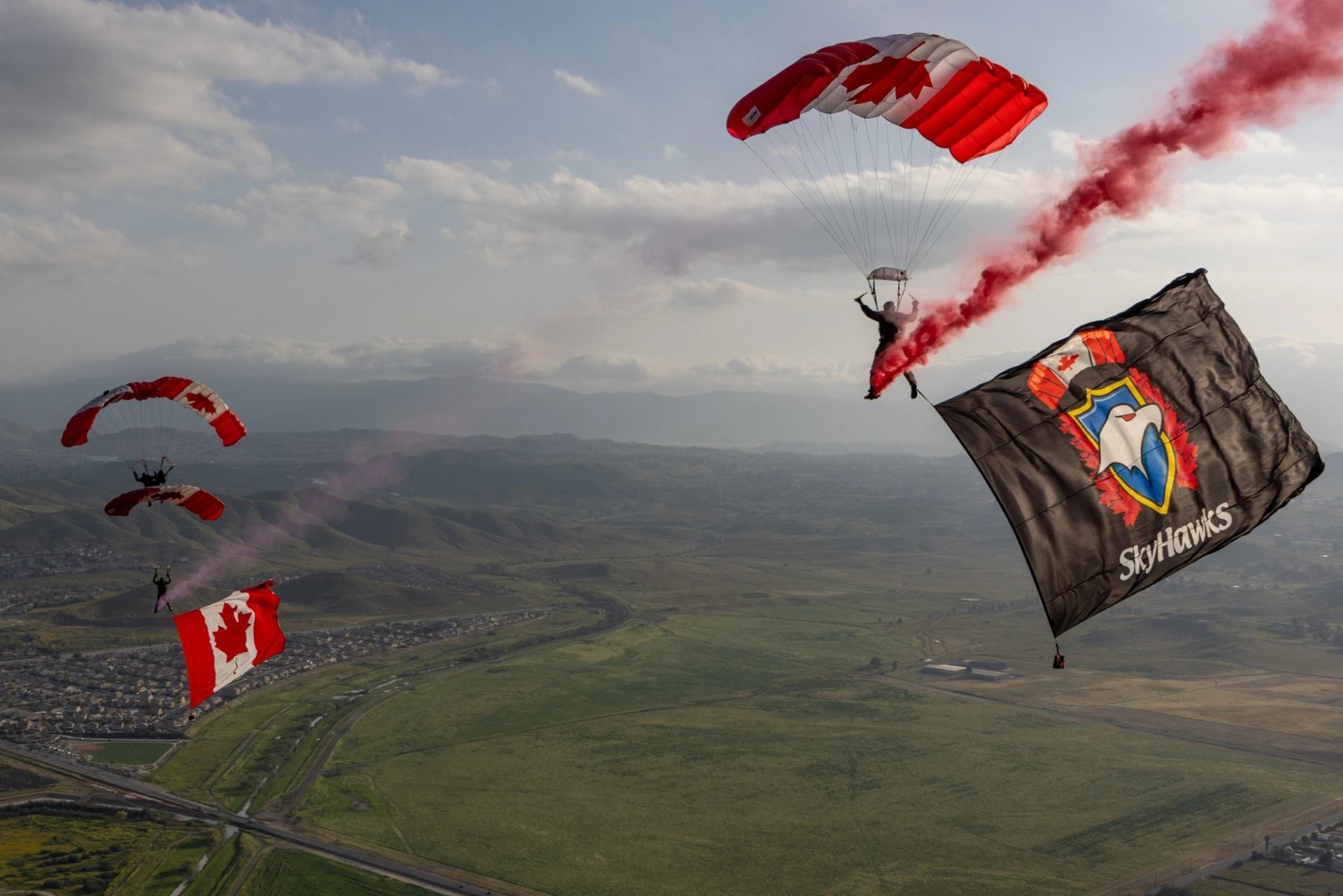 Parachutists with Canadian flag canopies trail smoke and large SkyHawks flag over landscape.