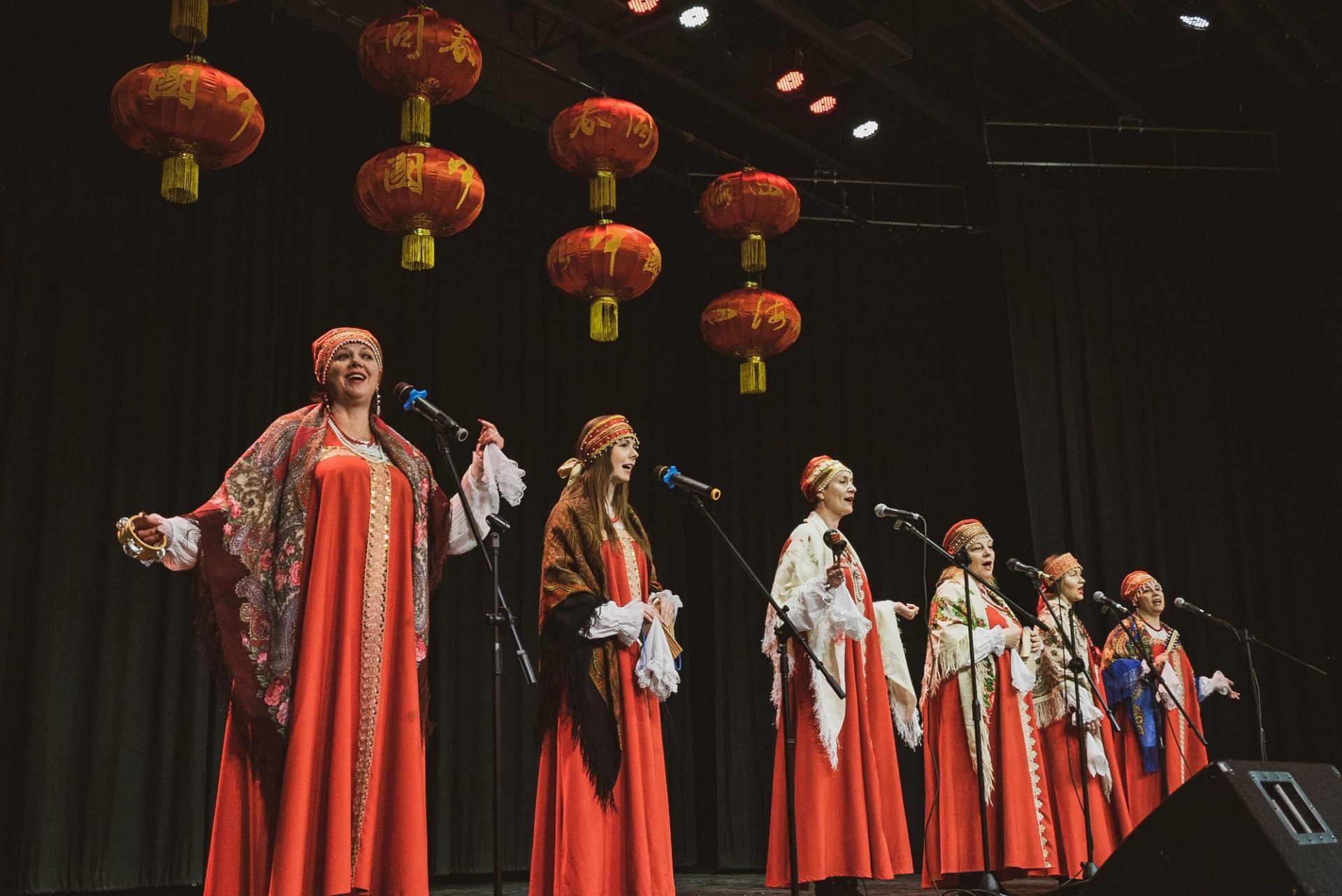 Group of performers in red robes singing on stage under hanging Chinese lanterns.