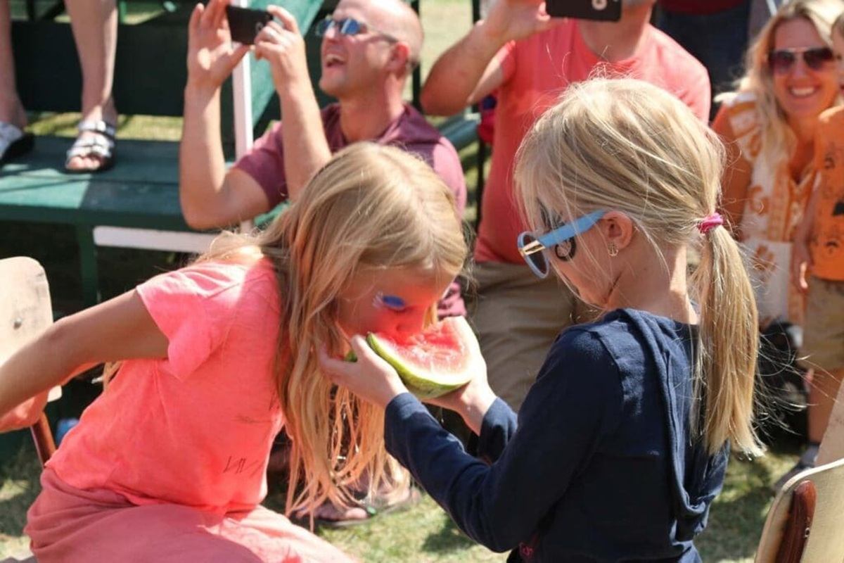 Children at outdoor event eat watermelon slice while adults watch and take photos.