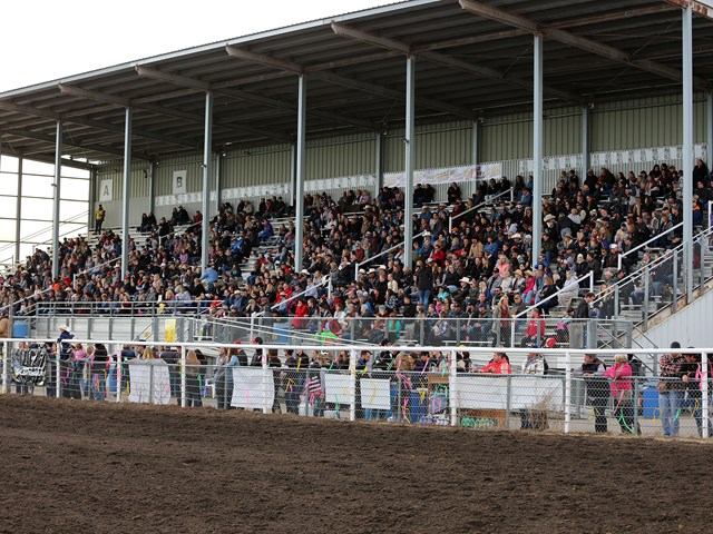 Grandstand crowd fills metal bleachers beside a fenced dirt arena at outdoor event.