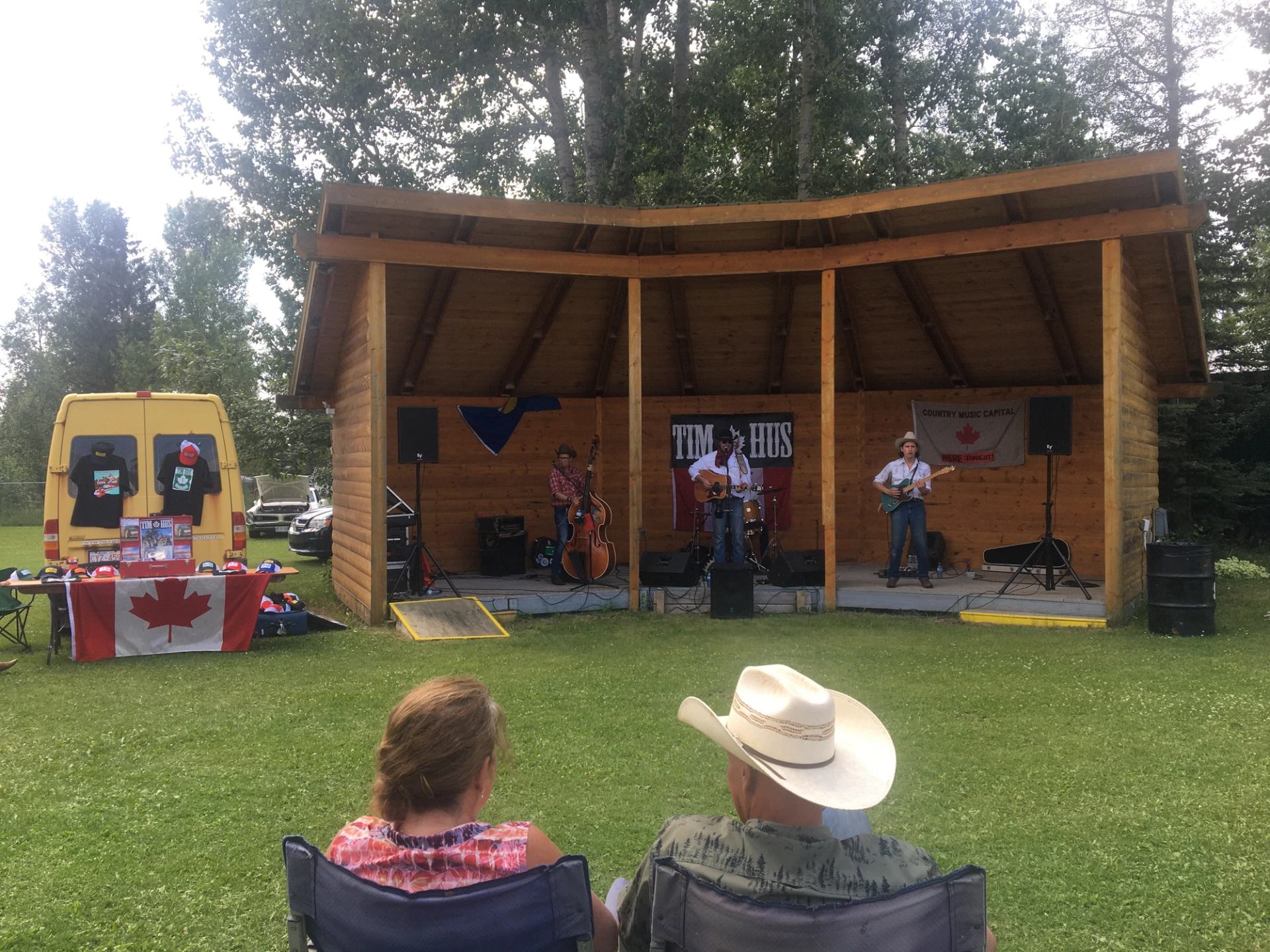 Outdoor band performs on wooden stage as seated audience watches, with Canadian flag merchandise.