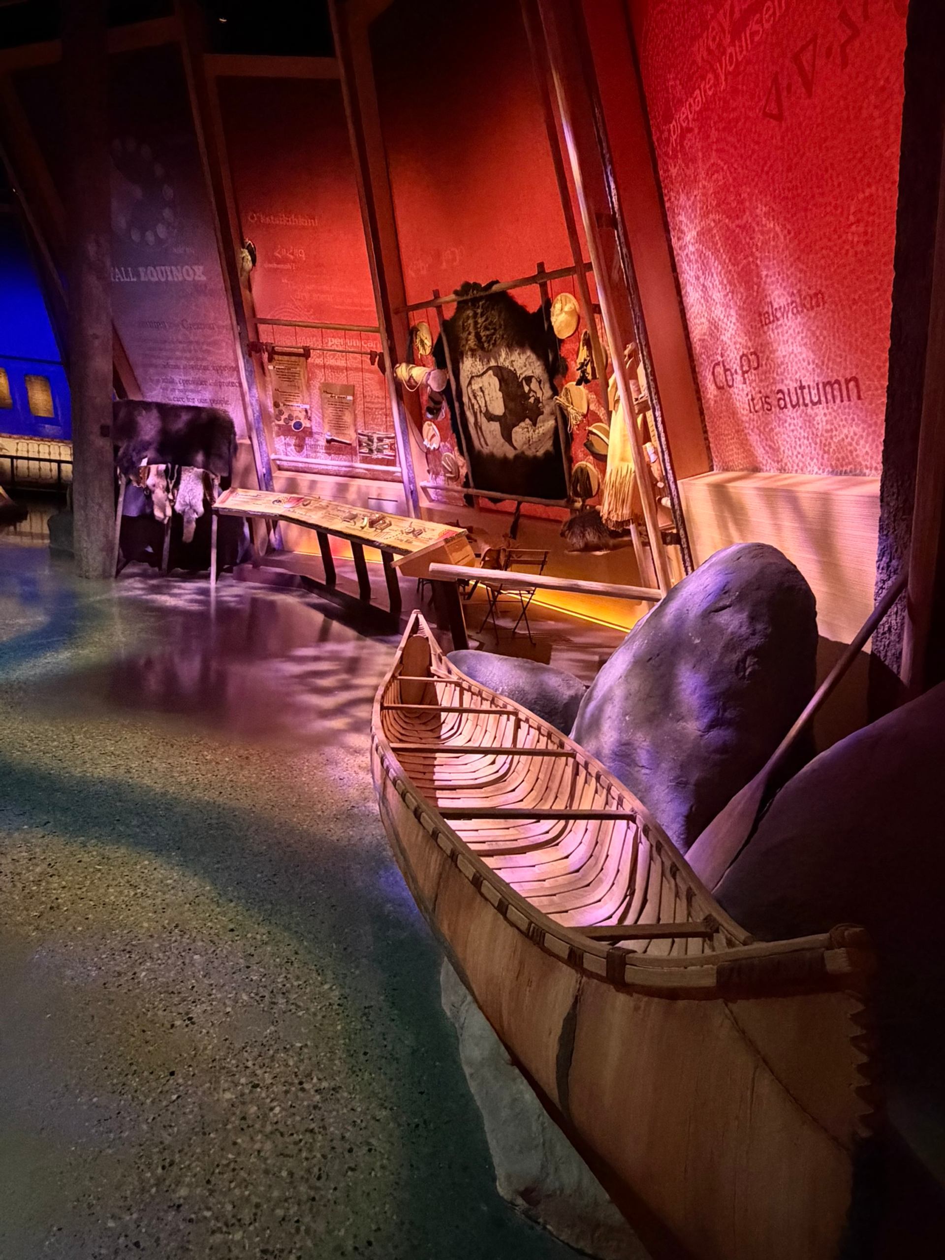 Museum exhibit with wooden canoe, rocks, animal hides and illuminated interpretive panels.