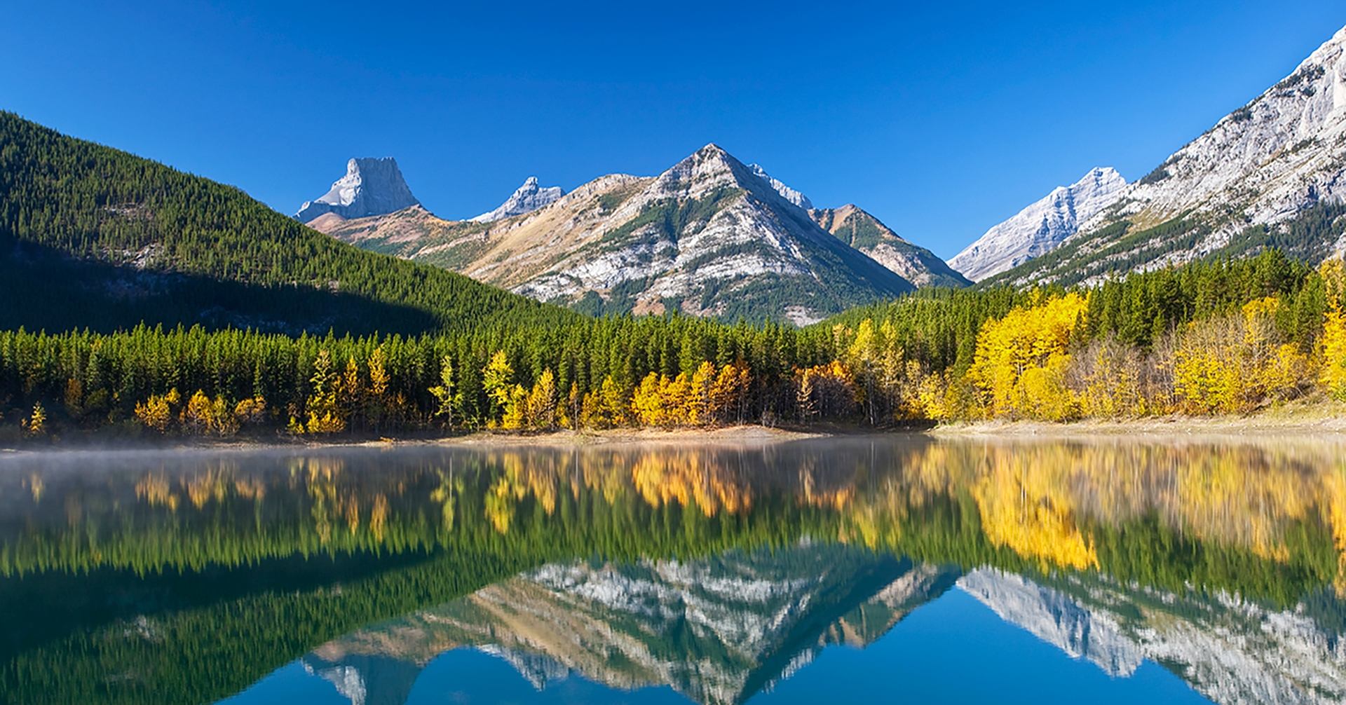 Crystal-clear lake reflecting autumn trees and rugged mountain peaks in Bow Valley.