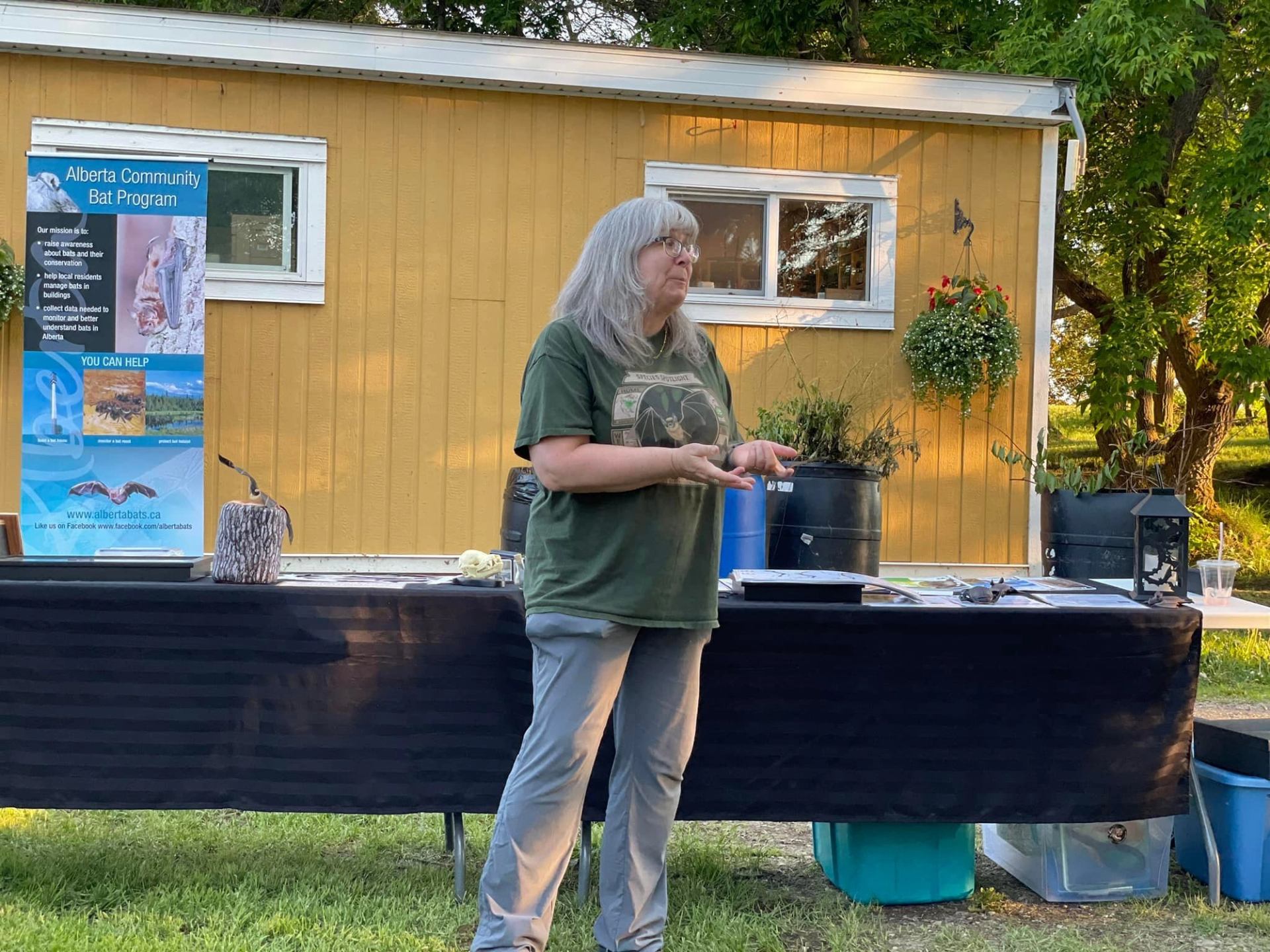 Presenter stands by Alberta Community Bat Program display table outside yellow shed with trees.