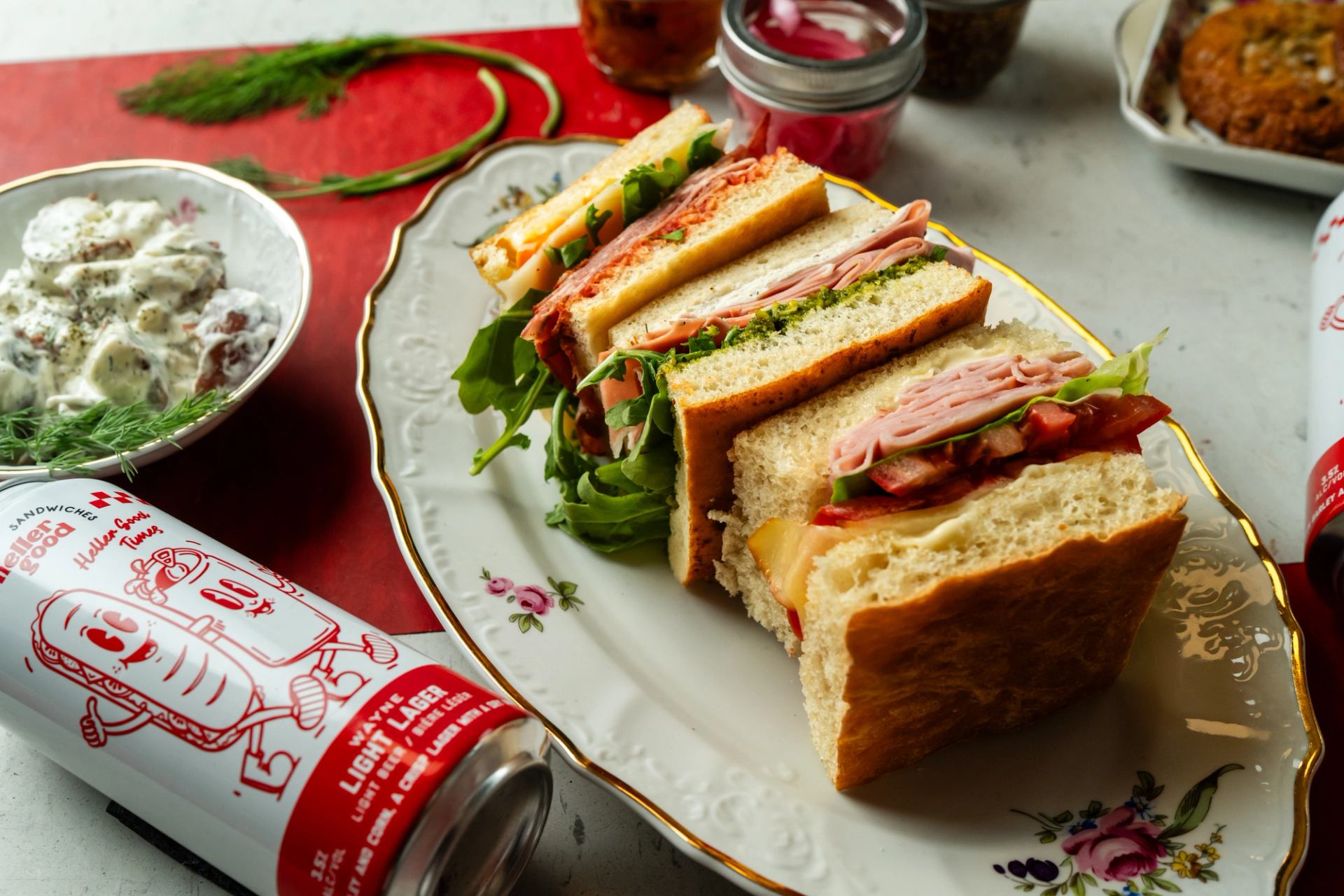 A plate of various deli sandwiches, a bowl of potato salad, and a "Better Good Light Lager" can on a red surface.