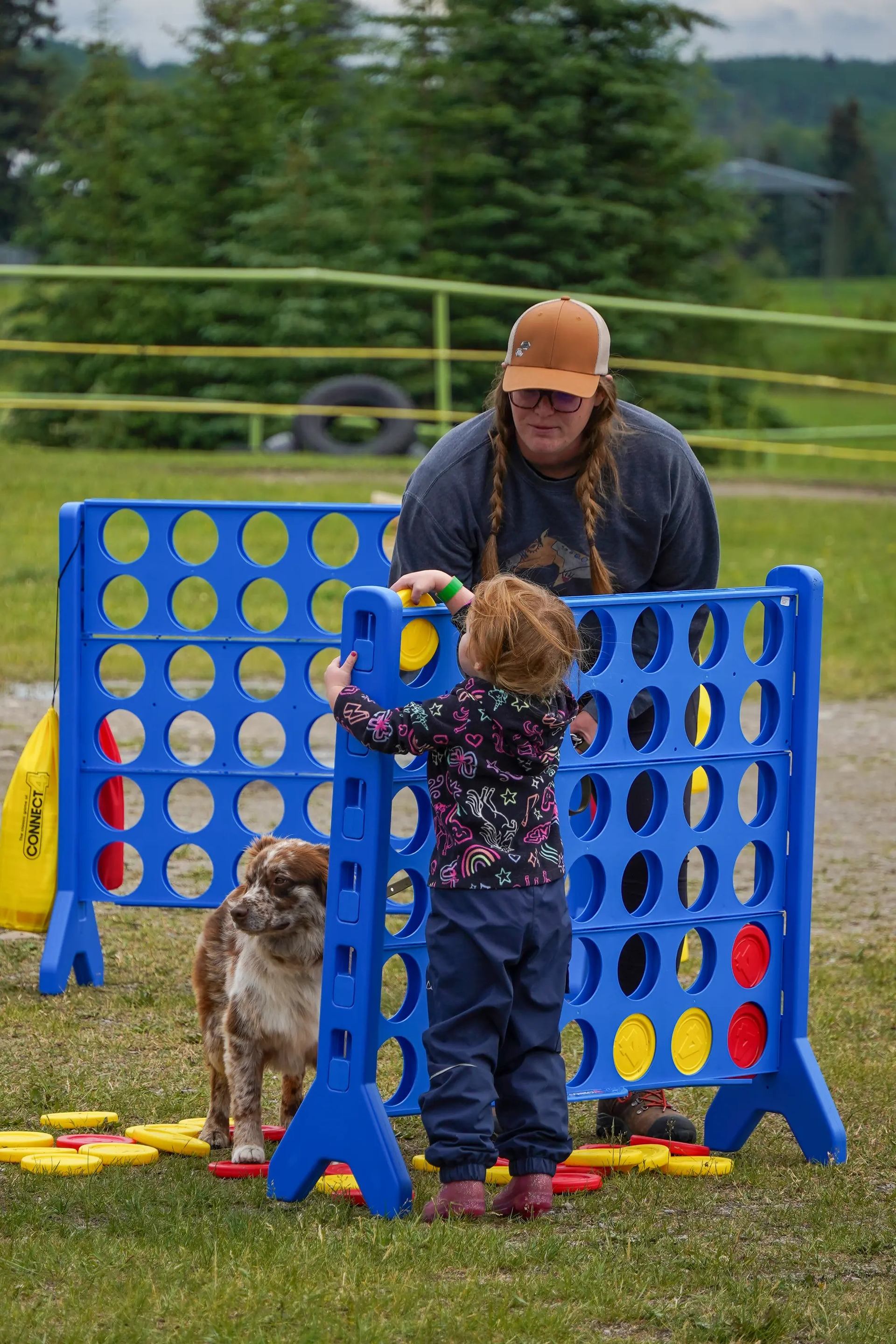 Child and adult play giant Connect Four outdoors with a dog nearby.