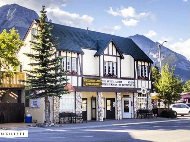 White Waterton Lakes Opera House building with sign and trees, in front of mountain peaks.