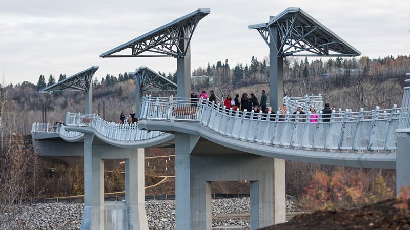 Curved pedestrian bridge with overhead canopies and people walking above a wooded river valley.