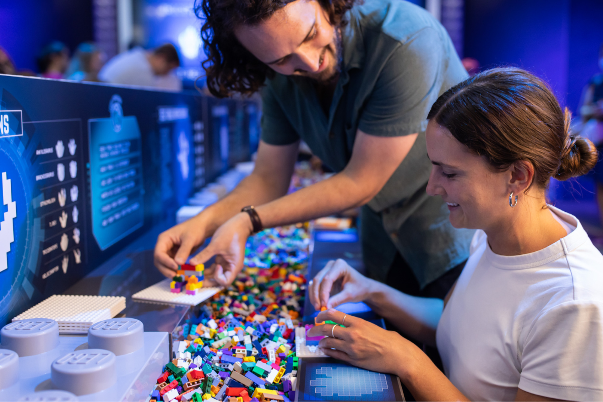 Two people build with LEGO bricks at a table in an interactive Jurassic World exhibit.