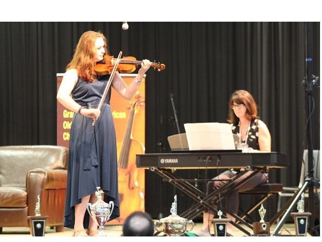 Violinist performs on stage beside pianist at keyboard, with trophies displayed in front.