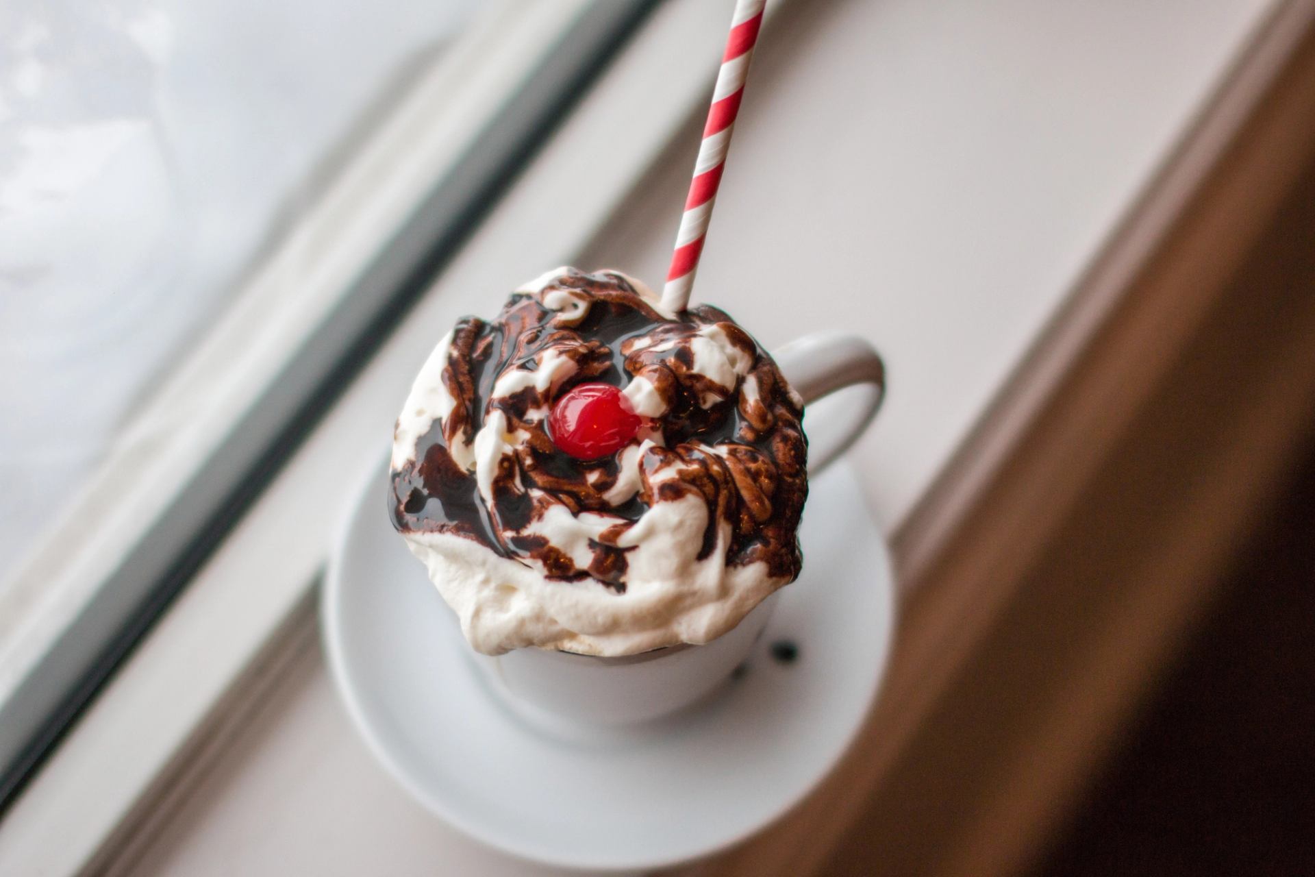 Hot chocolate with whipped cream, syrup, cherry, and striped straw by a window.