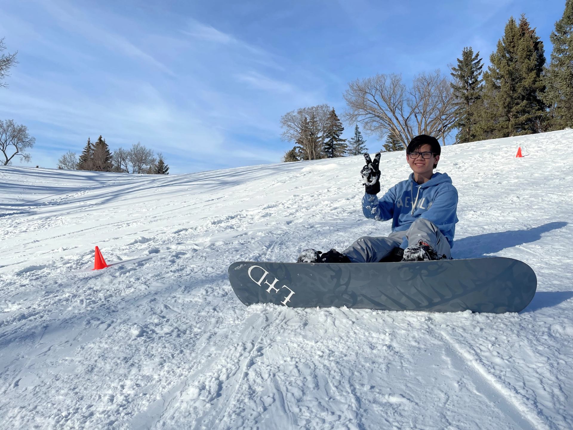 Snowboarder sitting on snow holding a snowboard on a ski slope with orange cone markers.