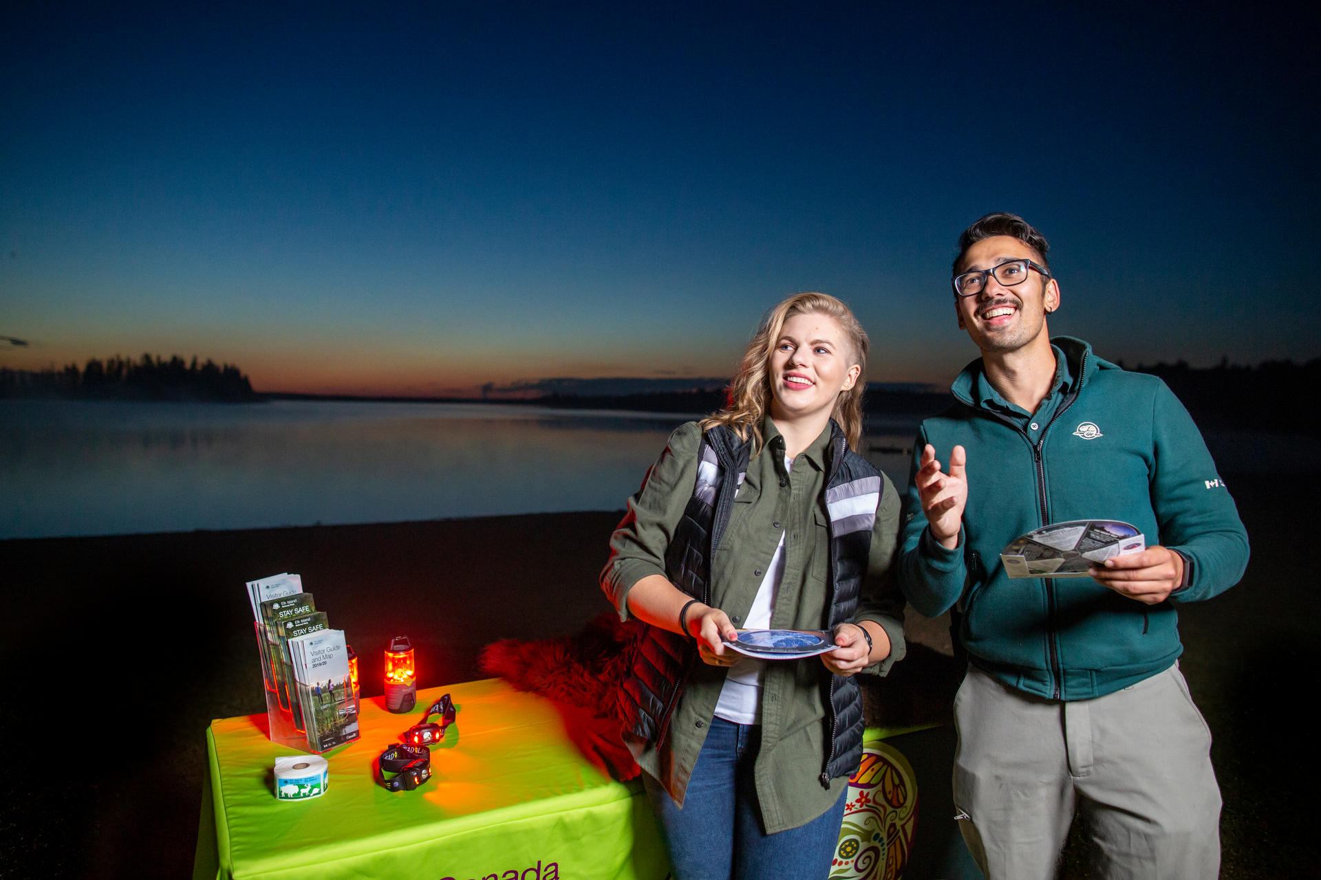 Two people hold maps beside a lit information table overlooking a lake.