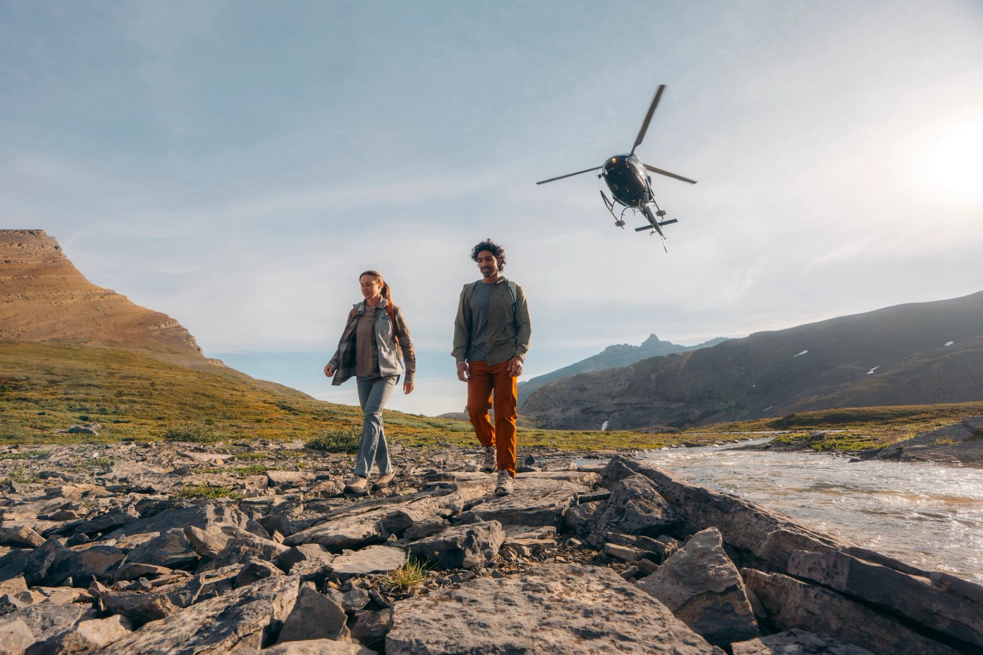 Two people hiking through a rocky mountain valley with a helicopter flying above.