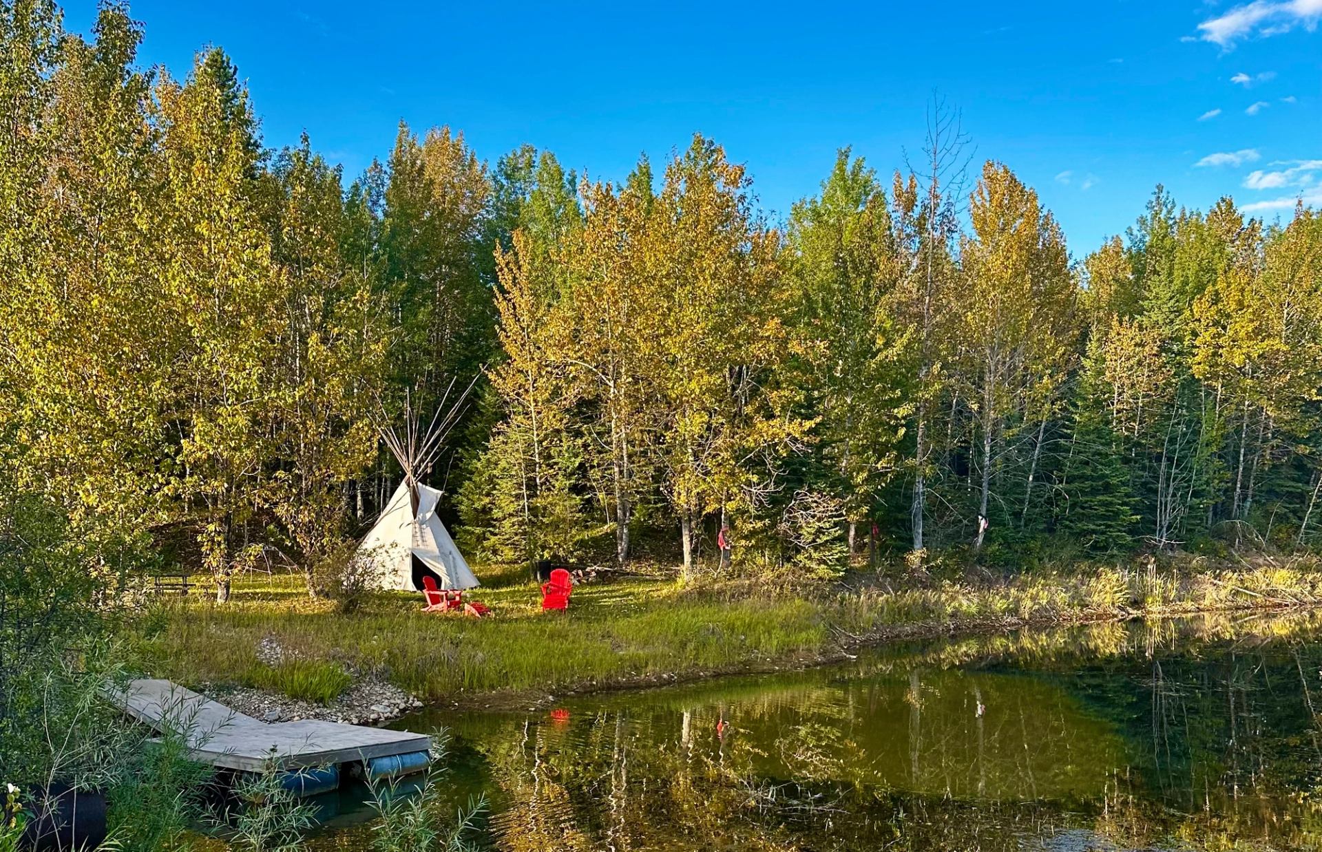 Tipi and red chairs beside a pond surrounded by green and yellow trees.