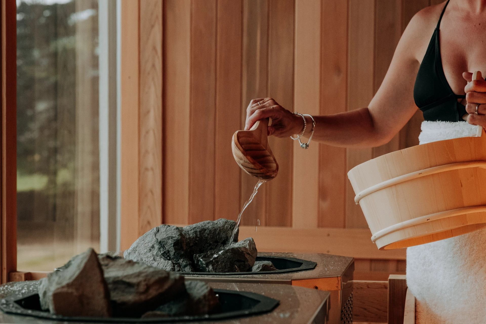 Person pouring water on sauna rocks in a wood-paneled steam room.
