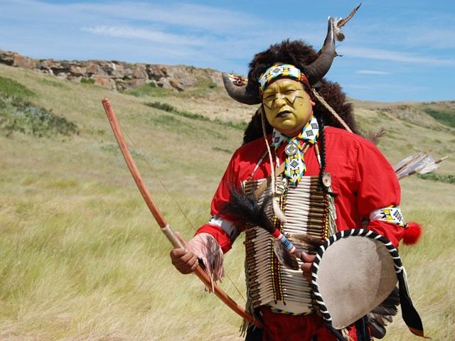 Native attire with bow, shield, and headdress in nature.