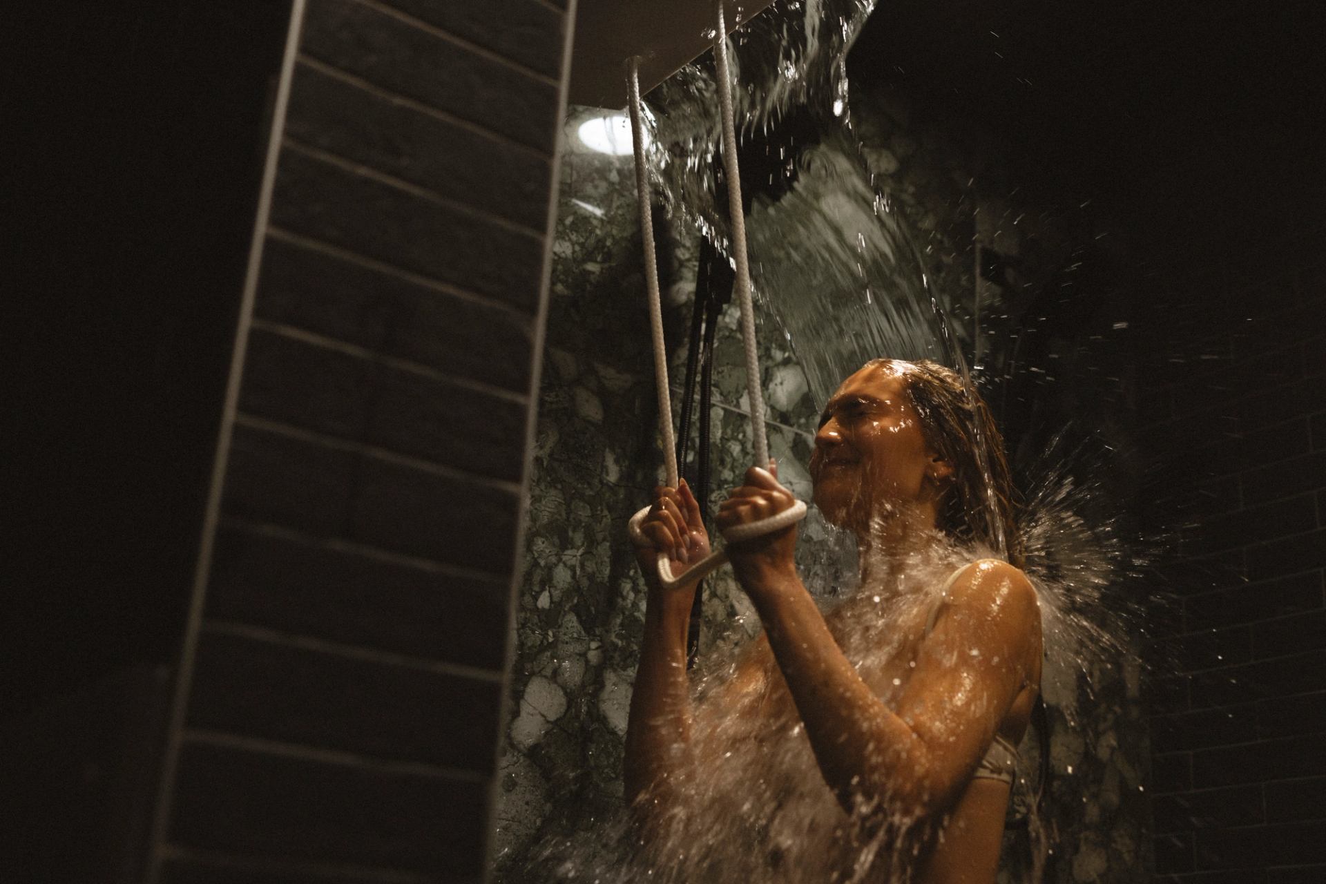 Person standing under heavy shower stream, holding two hanging ropes in dim bathroom.