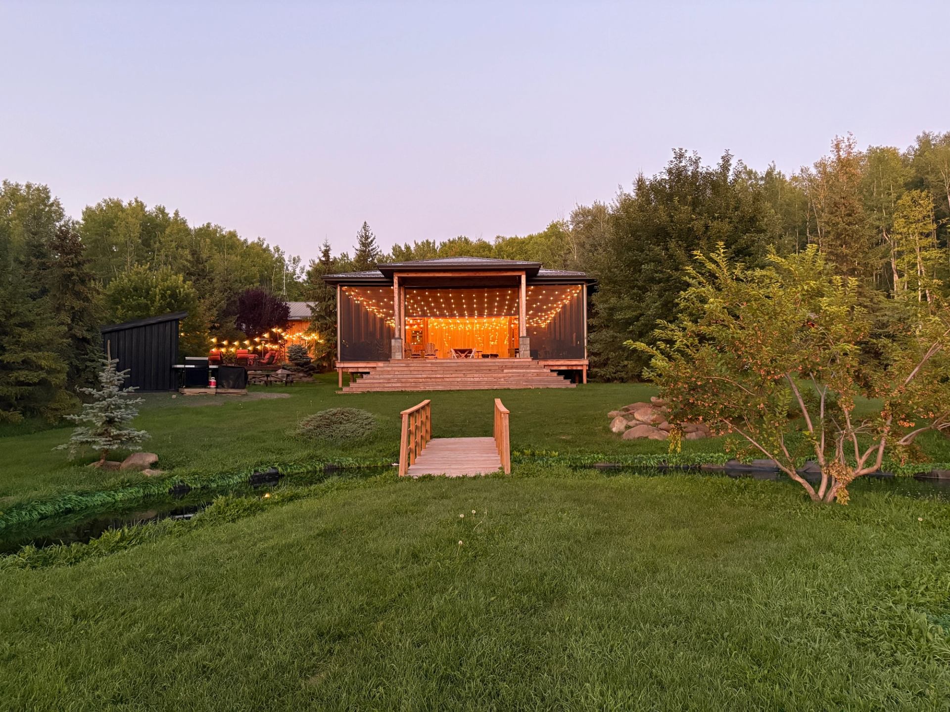 Cozy lodge lit with warm lights at dusk, surrounded by trees and open green lawn.