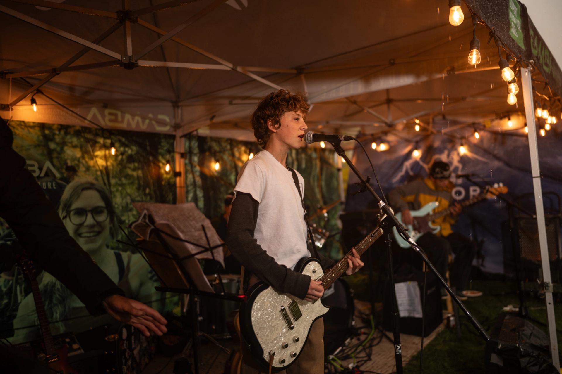 Guitarist sings into microphone under canopy with string lights and band equipment behind.