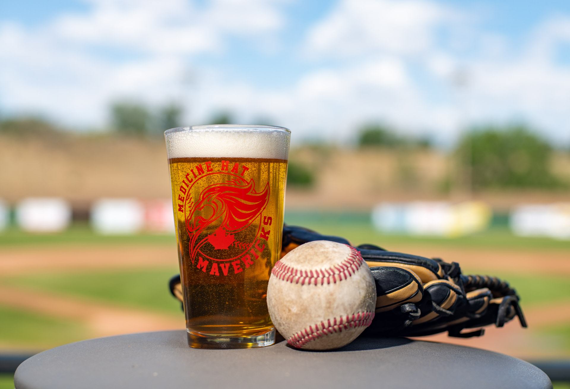 A pint glass with the Medicine Hat Mavericks logo sits next to a baseball and glove, with a blurred baseball field in the background.