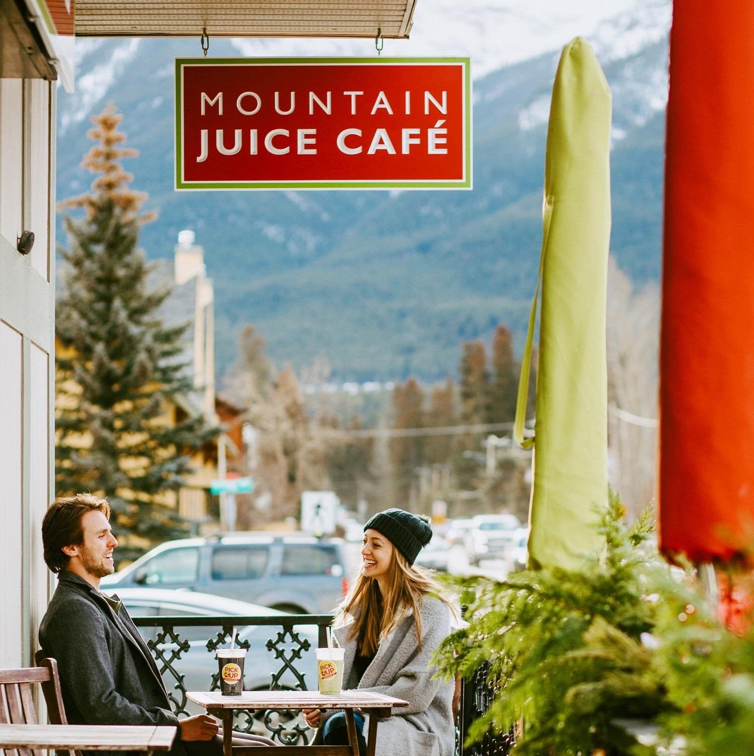 Two people at outdoor table under Mountain Juice Café sign with mountains and trees in background.
