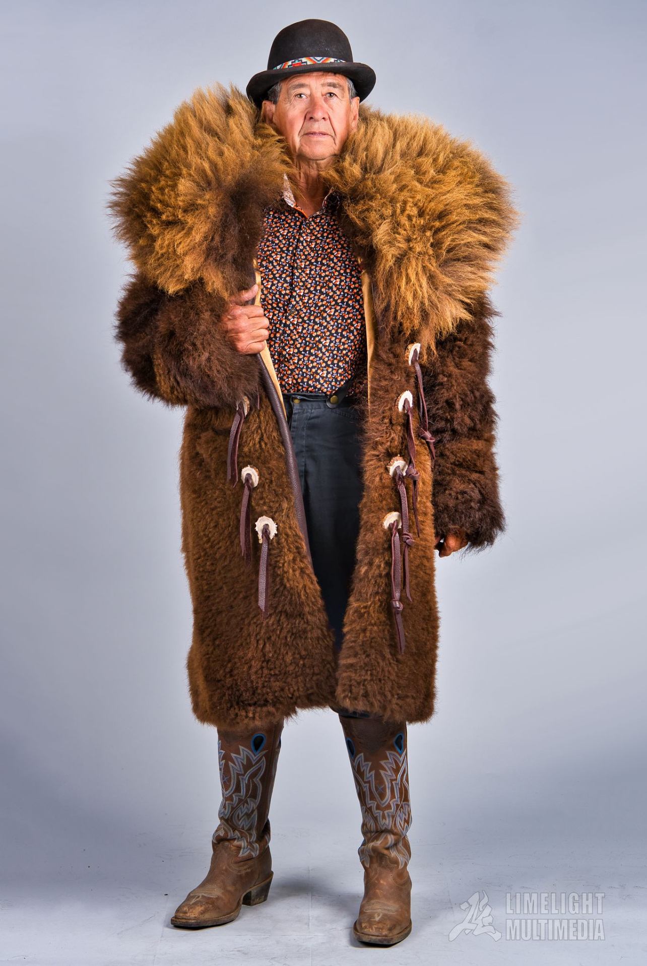 Person wearing long fur coat, patterned shirt, cowboy boots and hat against plain backdrop.