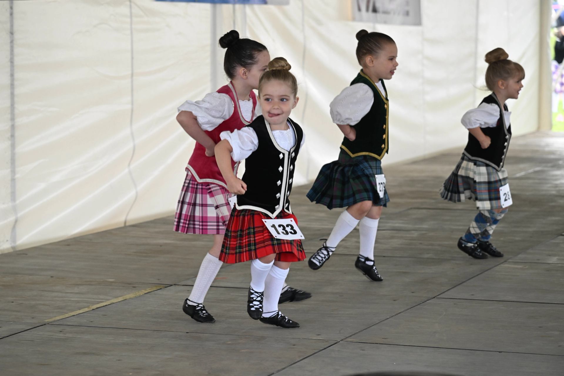 Children in kilts perform traditional Scottish dance indoors.