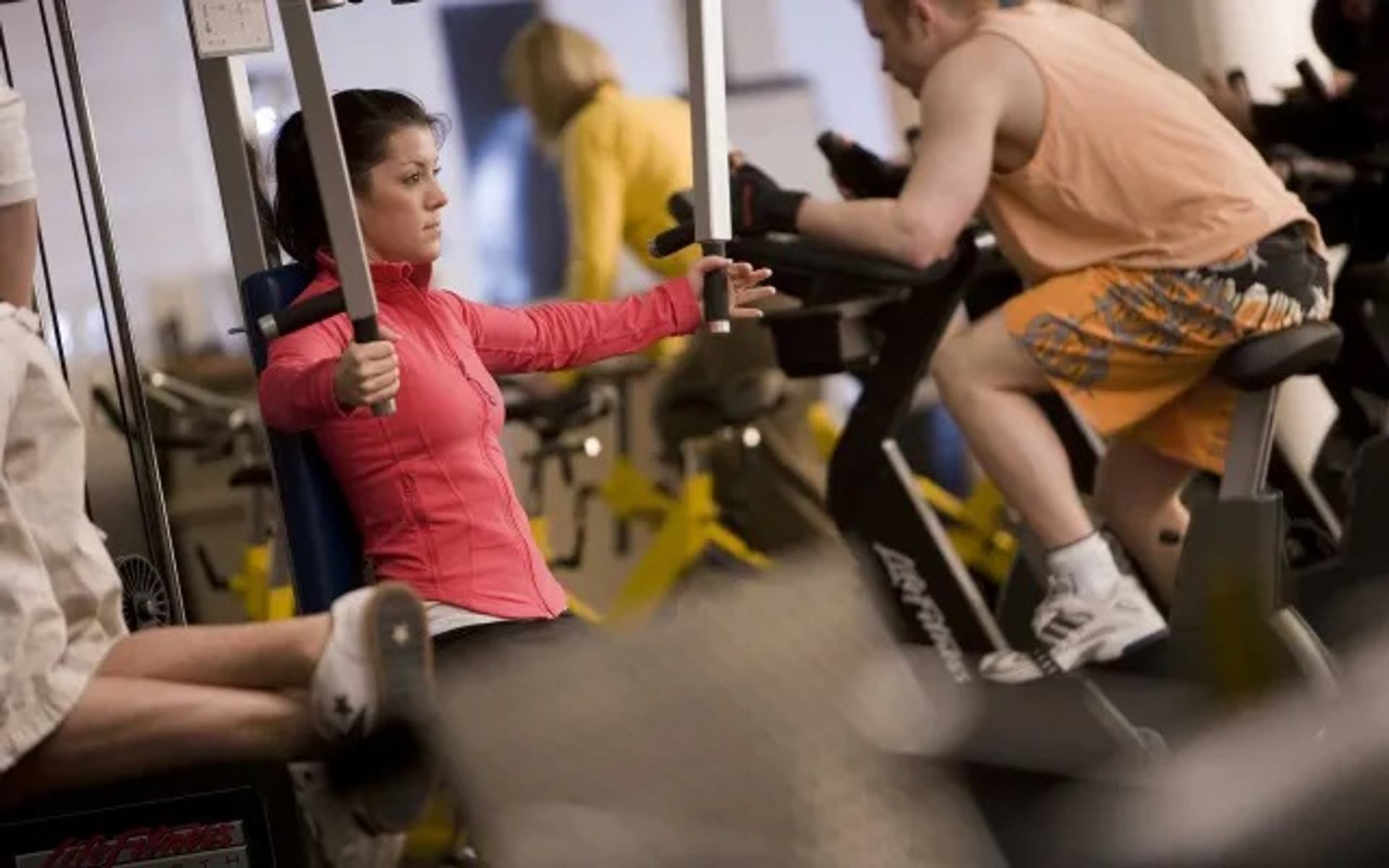A gym member uses a chest fly machine while others exercise on nearby fitness equipment.