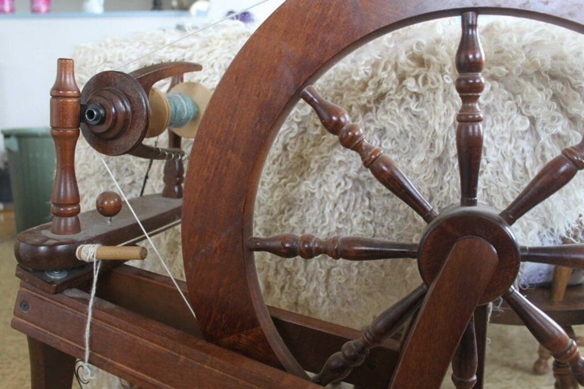 A wooden spinning wheel sits beside a large bundle of raw fleece, with thread being drawn onto the s