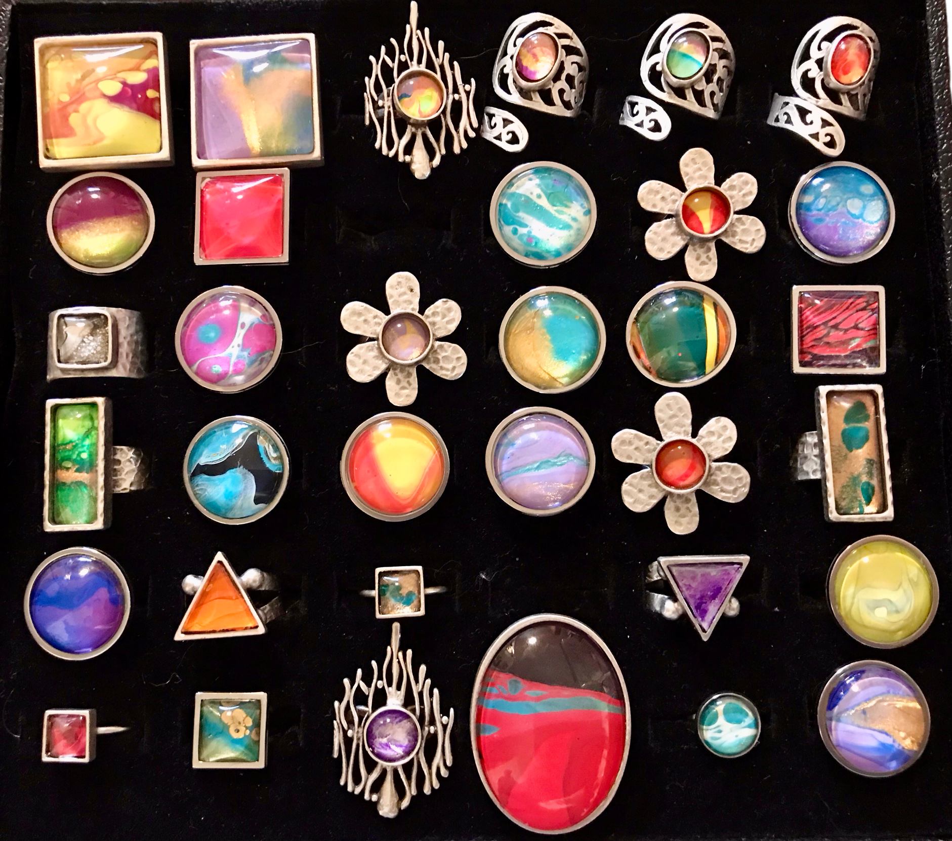 A collection of rings with colorful, abstract art designs in various shapes and metal settings, displayed on a black background.
