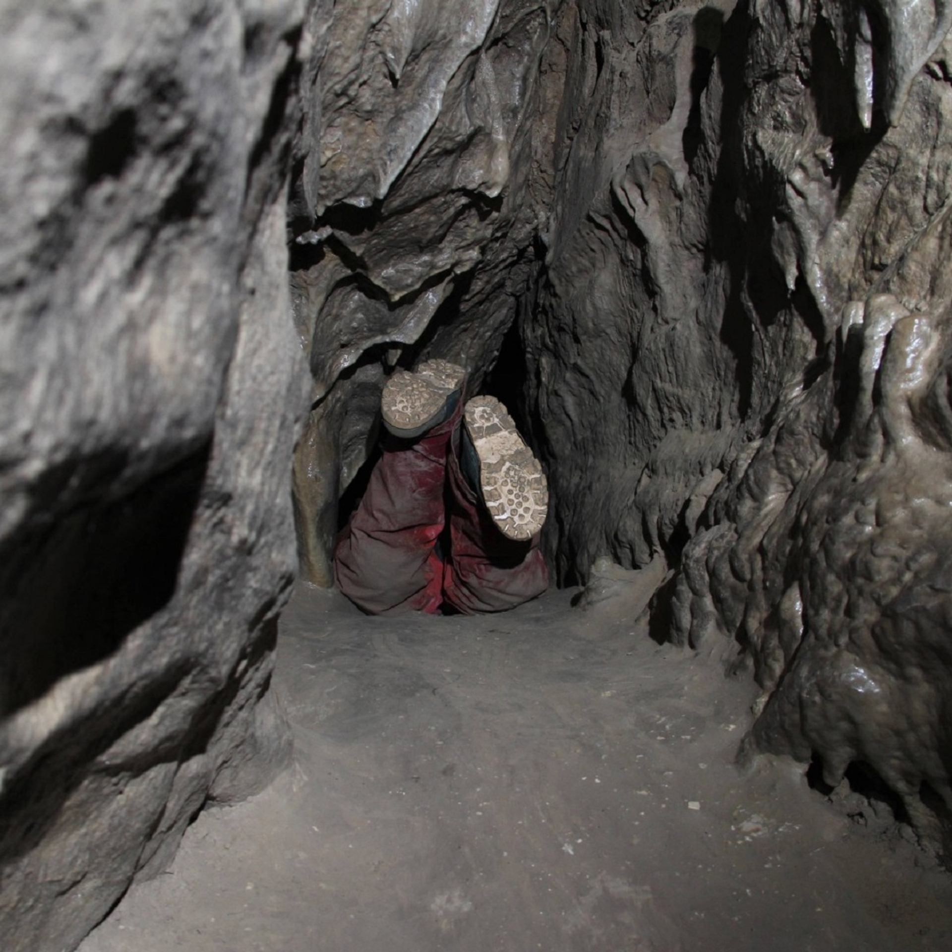 A person navigates a narrow, muddy squeeze passage inside Rat’s Nest Cave, surrounded by rough limestone walls.