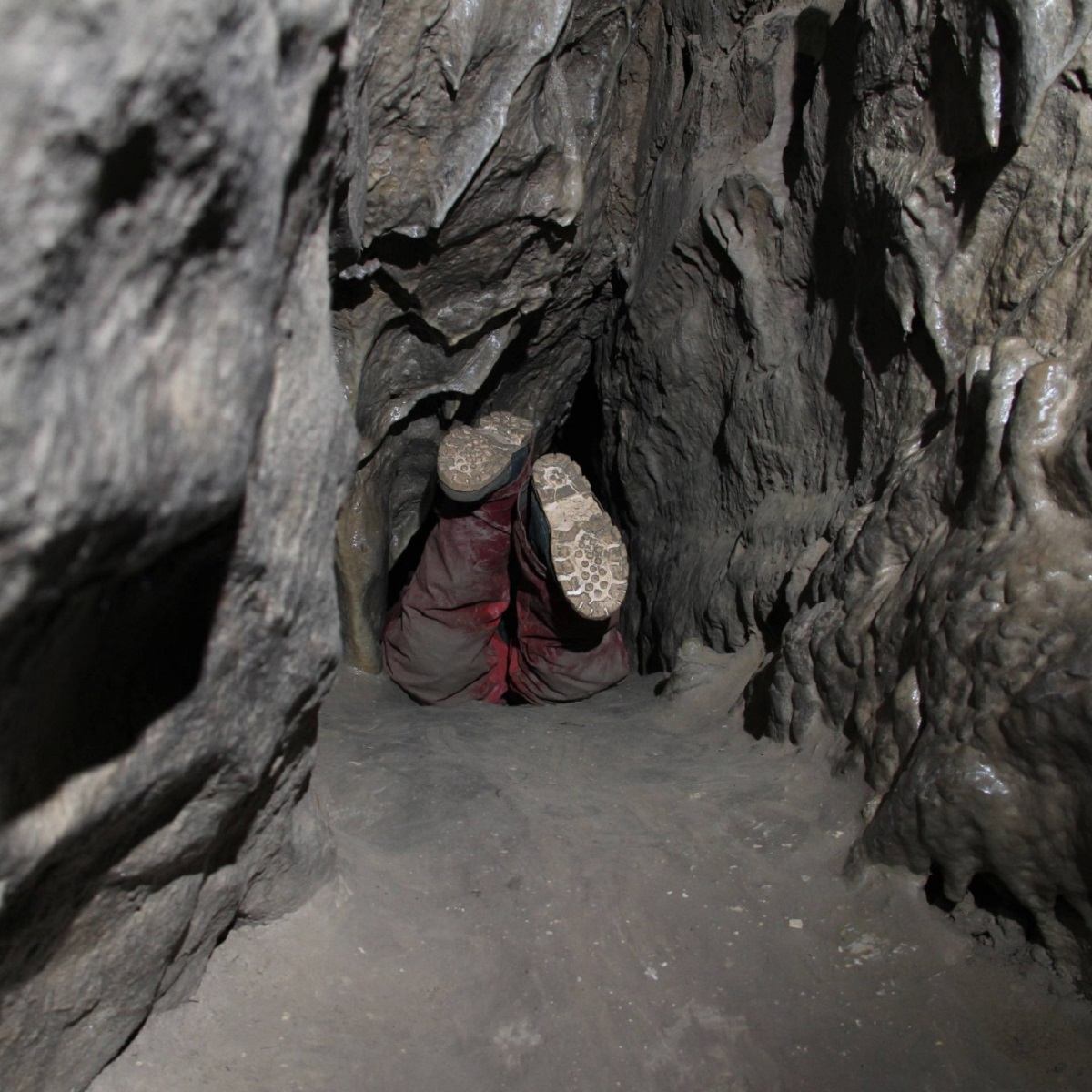 A person navigates a narrow, muddy squeeze passage inside Rat’s Nest Cave, surrounded by rough limestone walls.