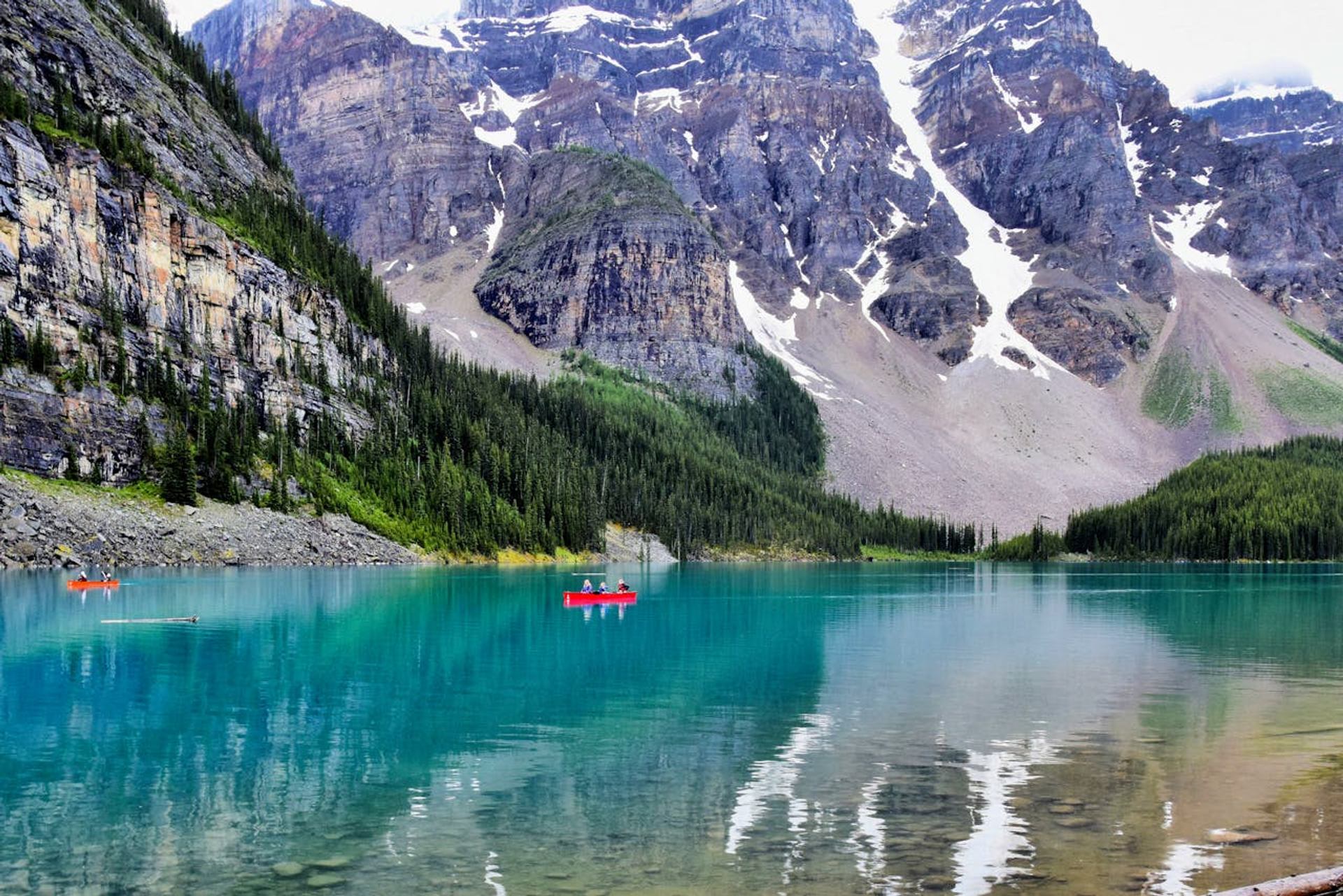 Turquoise mountain lake with red canoes, evergreen forest, and rocky peaks with snow patches.