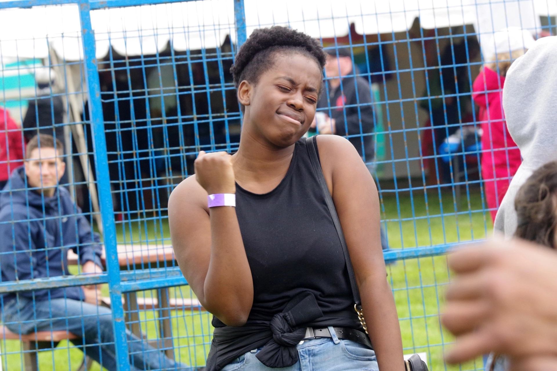 Festival attendee in black tank top with wristband, standing near blue metal fence.